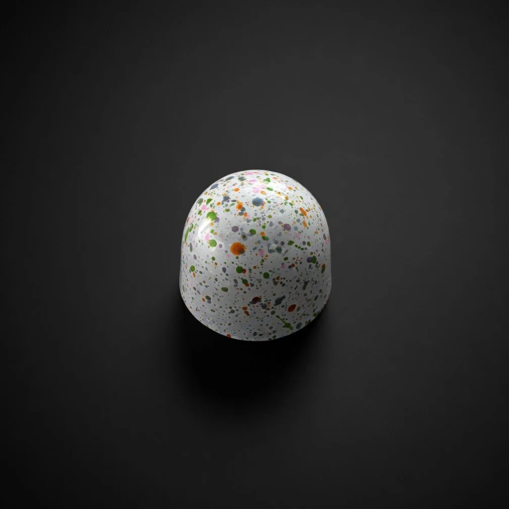 A glossy white chocolate bonbon covered in colorful speckles of pink, green, orange, and blue, sharply in focus and well lit, resting on a solid matte black background with realistic contact shadow.