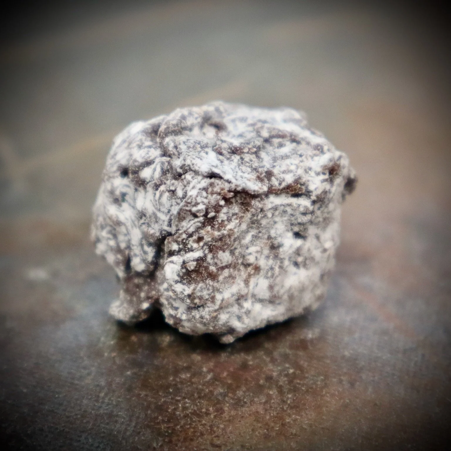 A close-up of a single chocolate truffle covered in powdered sugar on a dark surface.