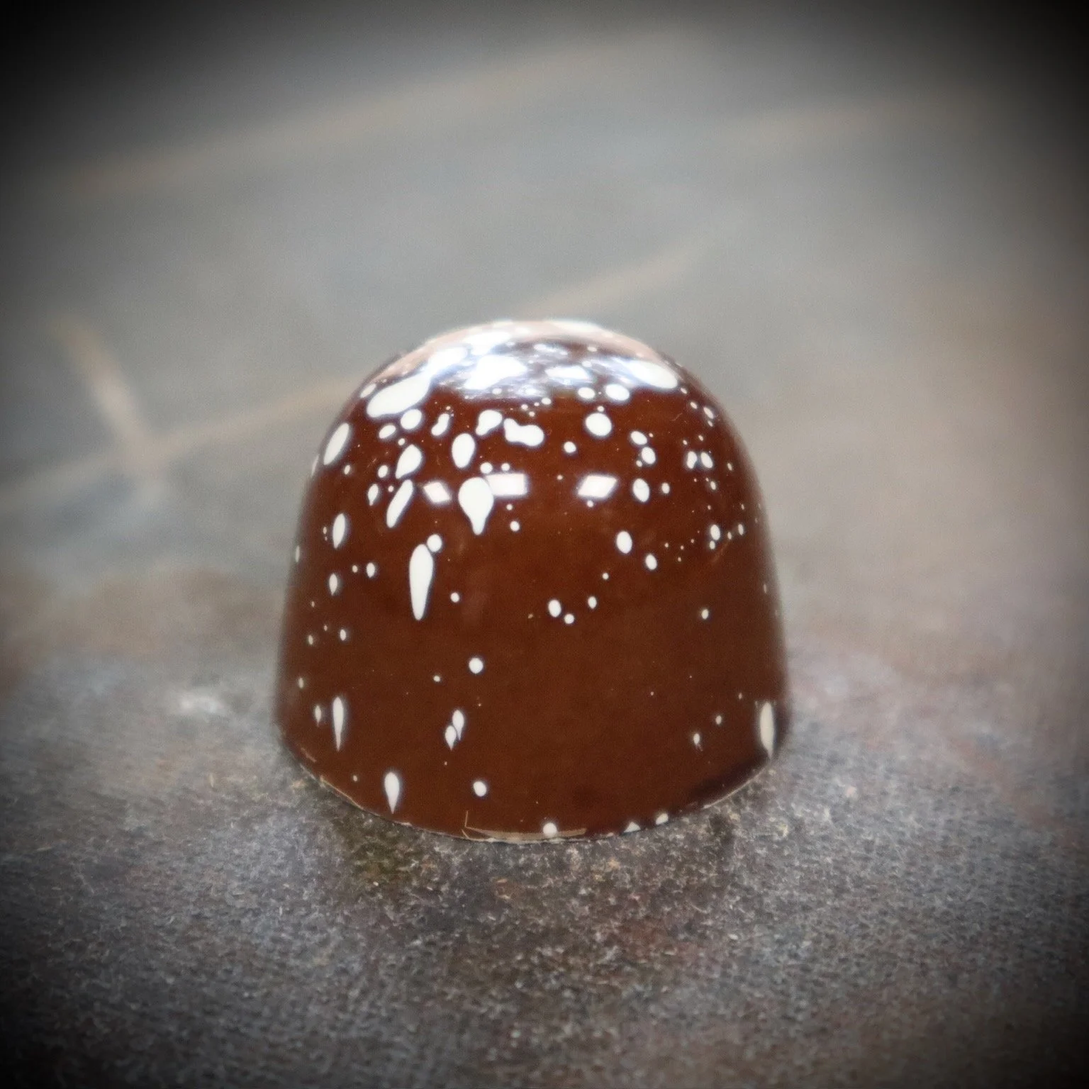 Close-up of a chocolate praline with white speckles on top, sitting on a dark surface.