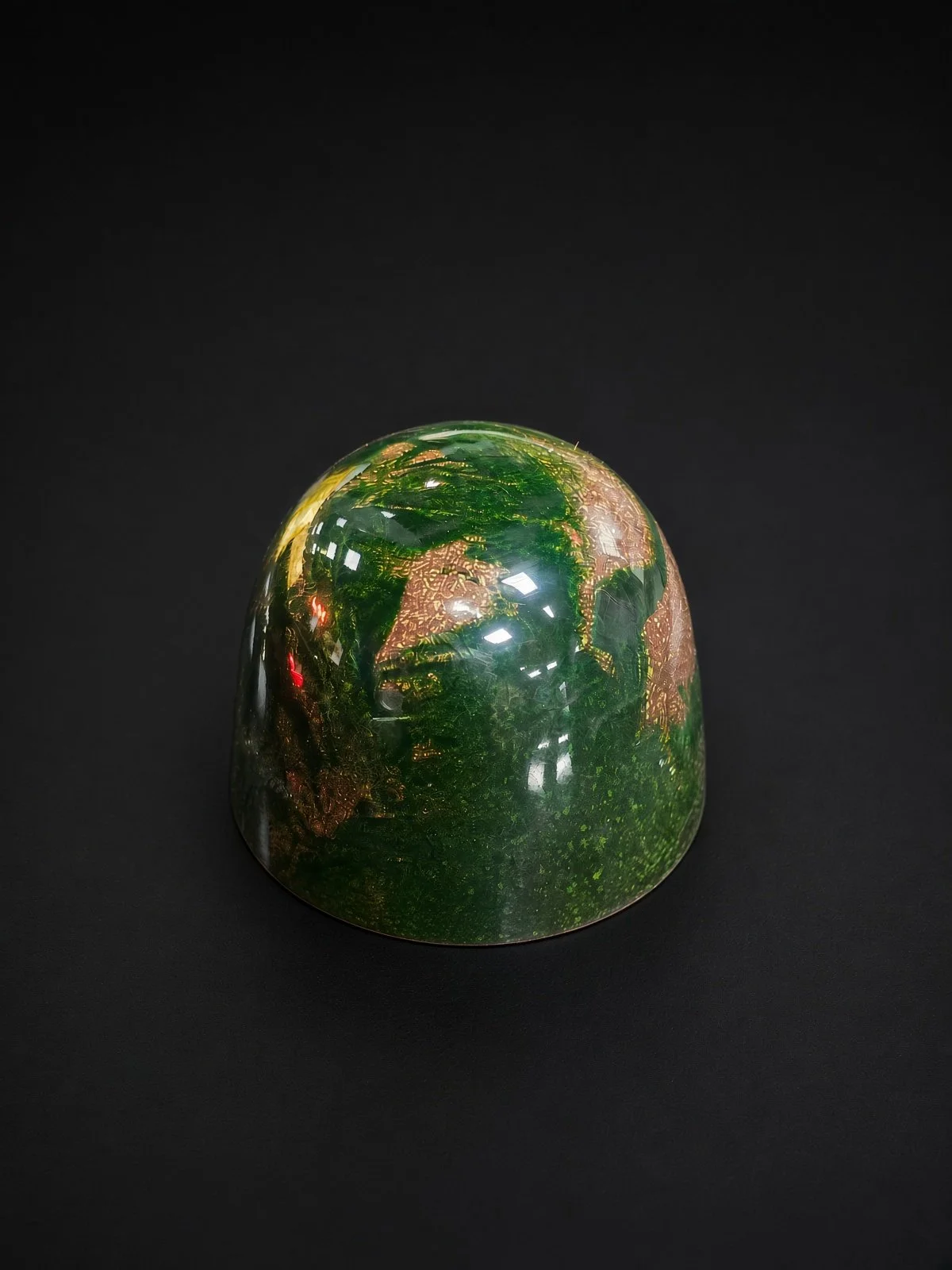 A glossy deep green chocolate bonbon with mossy, textured green patches and copper-bronze accents, sharply in focus and well lit, resting on a solid matte black background.