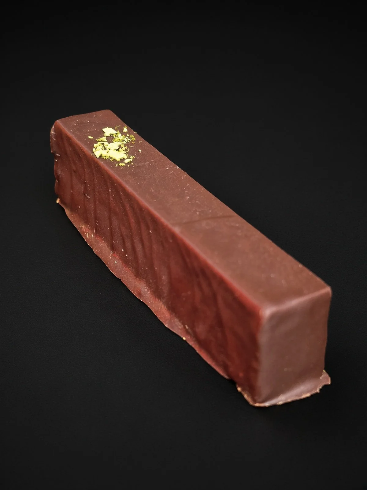 A long rectangular dark chocolate bar with a small dusting of green matcha on top, sharply in focus and well lit, resting on a solid matte black background.