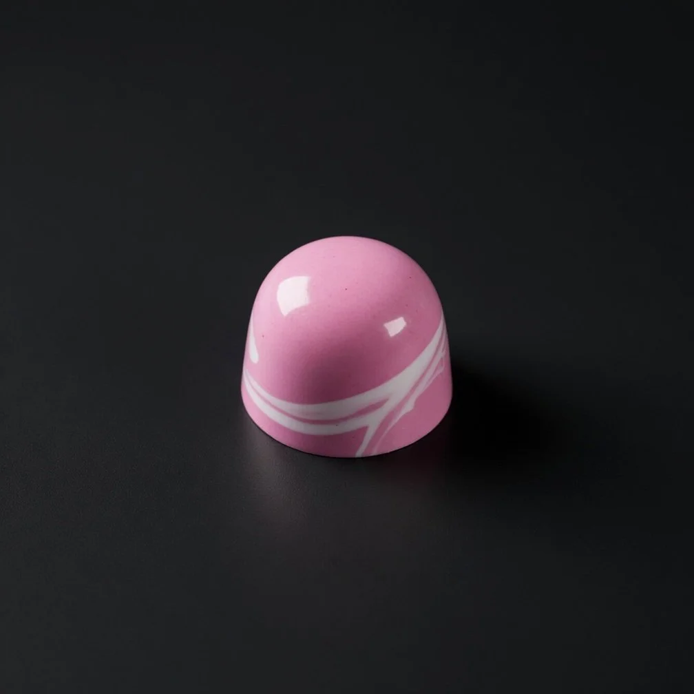 A glossy bright pink chocolate bonbon with elegant white marbled swirls, sharply in focus and well lit, resting on a solid matte black background with realistic contact shadow.