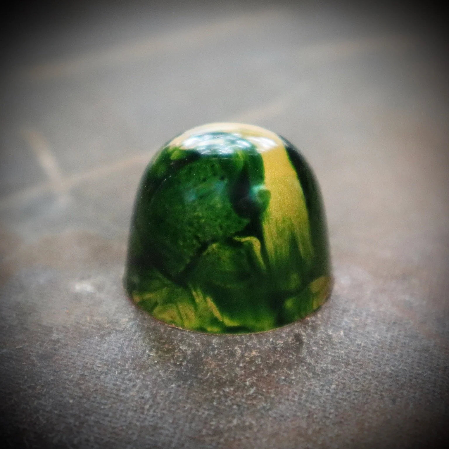 A green marble with yellow streaks on a textured gray surface.