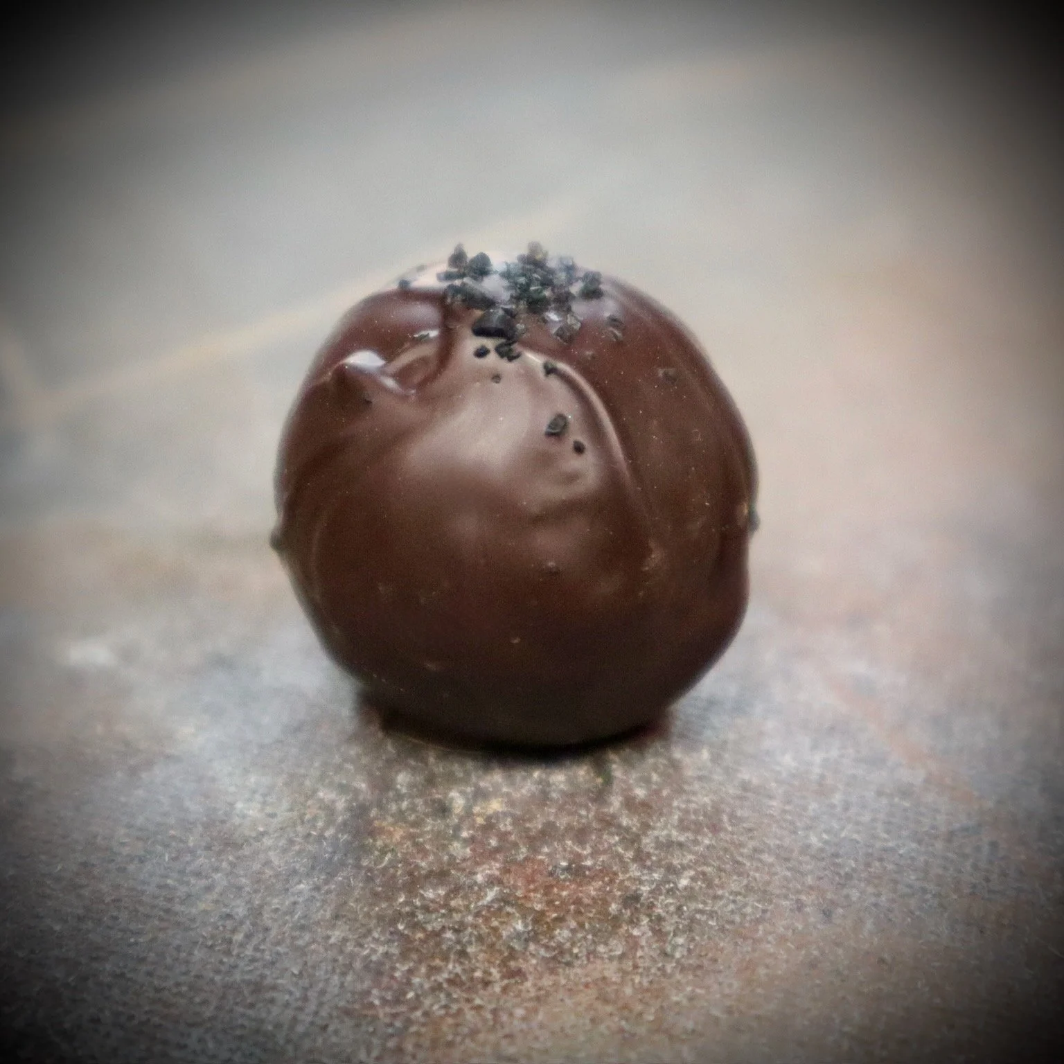 A small round chocolate truffle coated with a light dusting of cocoa powder and topped with a few sprinkles of sea salt.