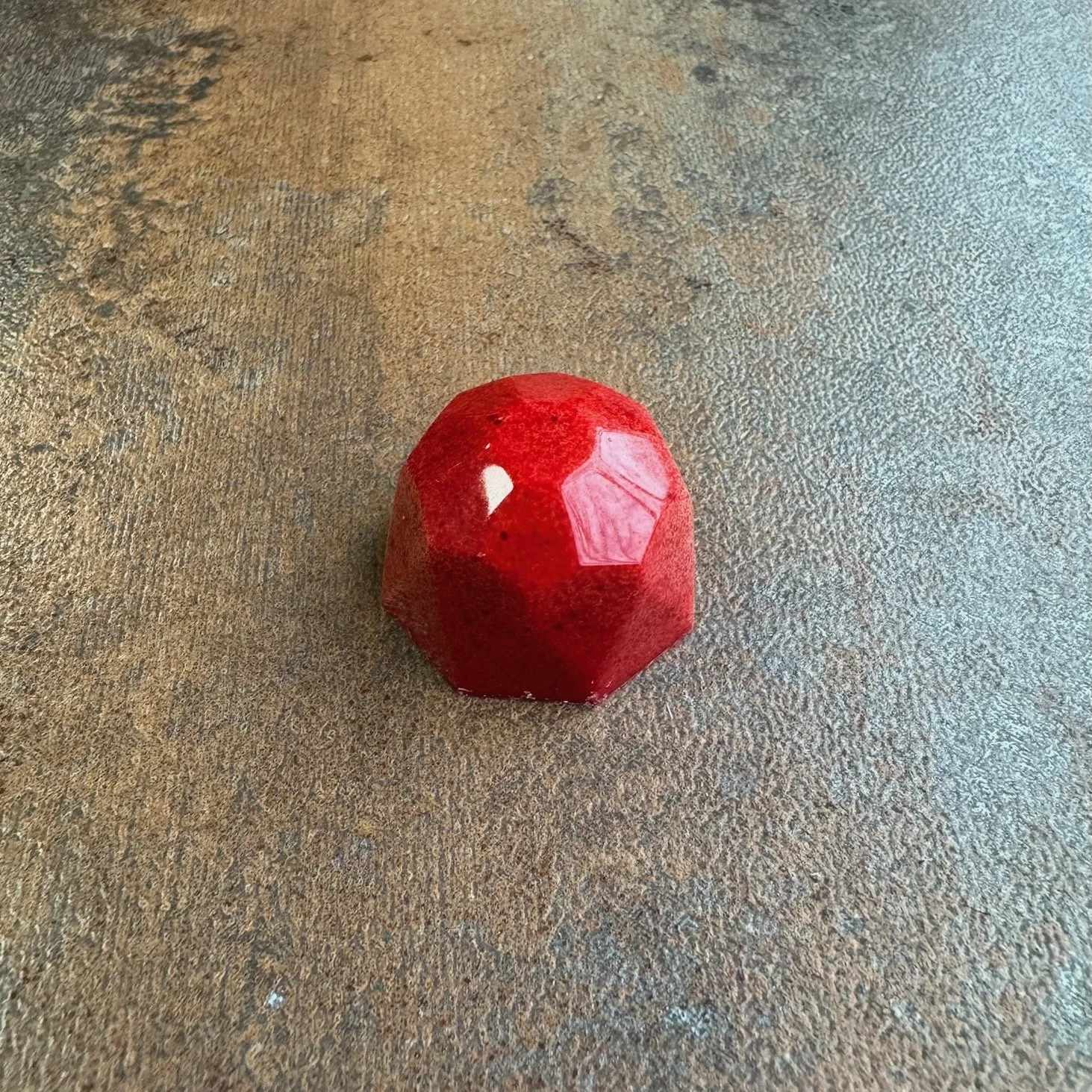A small, red, faceted object with a glossy surface, sitting on a dark surface.