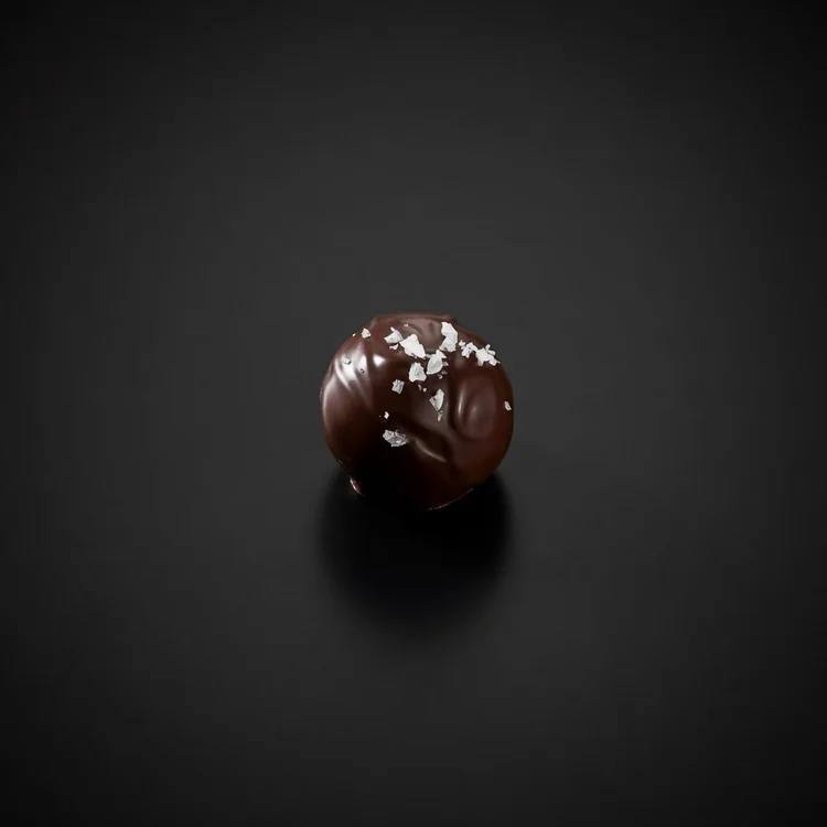A Goat Cheese Kinetic Bomb: a small glossy dark chocolate sphere topped with flakes of sea salt, resting on a solid matte black background.