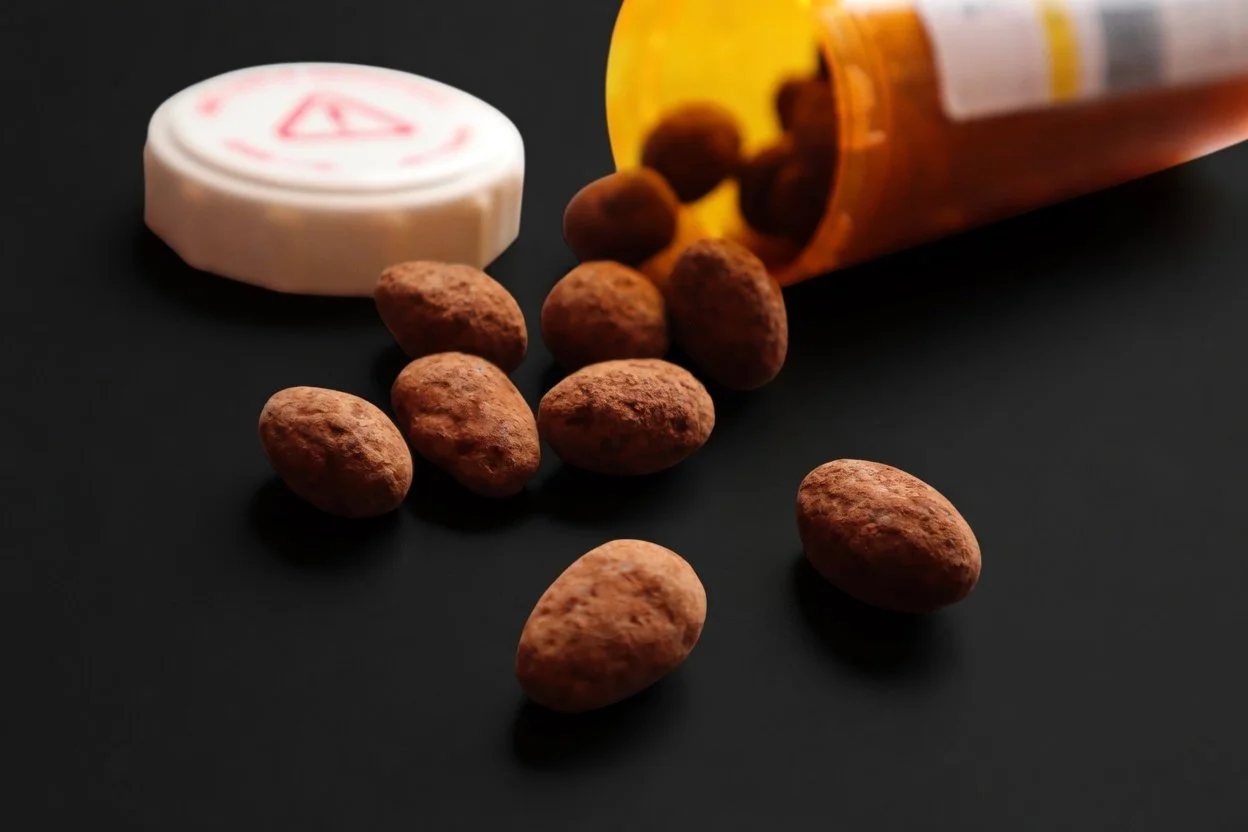 Chocolate covered almonds spilling out of an amber prescription bottle onto a black surface, with a white cap in the background.