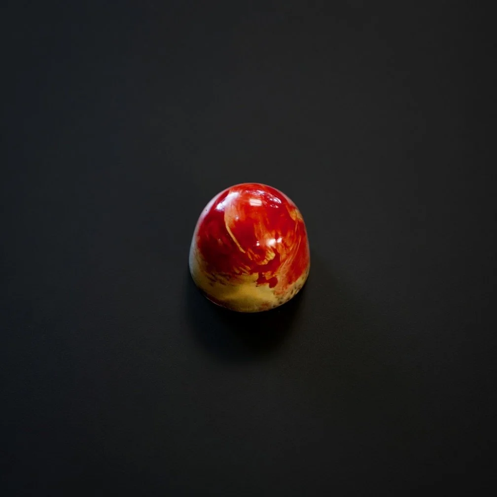 A glossy red-and-yellow marbled chocolate bonbon with vibrant swirls, sharply in focus and well lit, resting on a solid matte black background with realistic contact shadow.