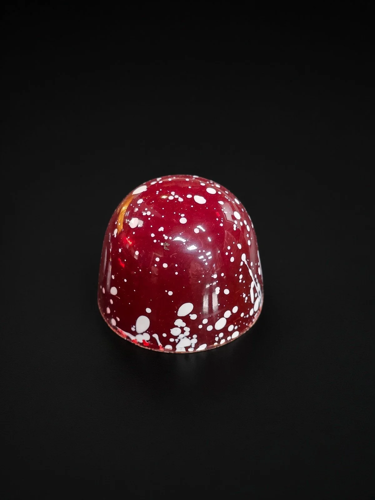 A glossy deep red dome-shaped chocolate bonbon speckled with white splatters, sharply in focus and well lit, resting on a solid matte black background.