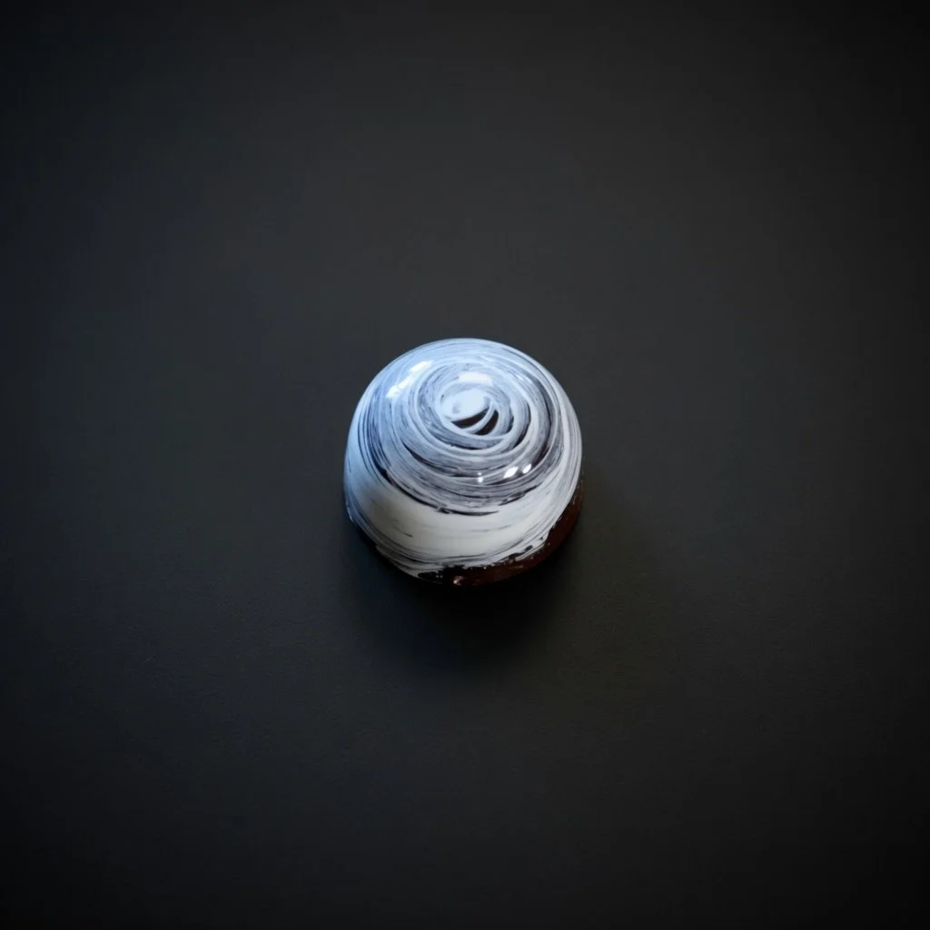 A glossy white-and-dark-blue swirled chocolate bonbon with hypnotic spiral pattern, sharply in focus and well lit, resting on a solid matte black background with realistic contact shadow.