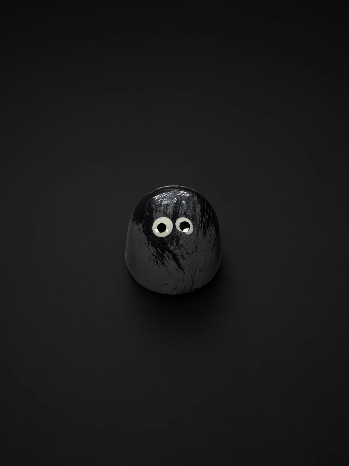 A glossy charcoal-gray dome-shaped chocolate bonbon with dark brushstroke streaks and two white candy eyes, sharply in focus and well lit, resting on a solid matte black background.