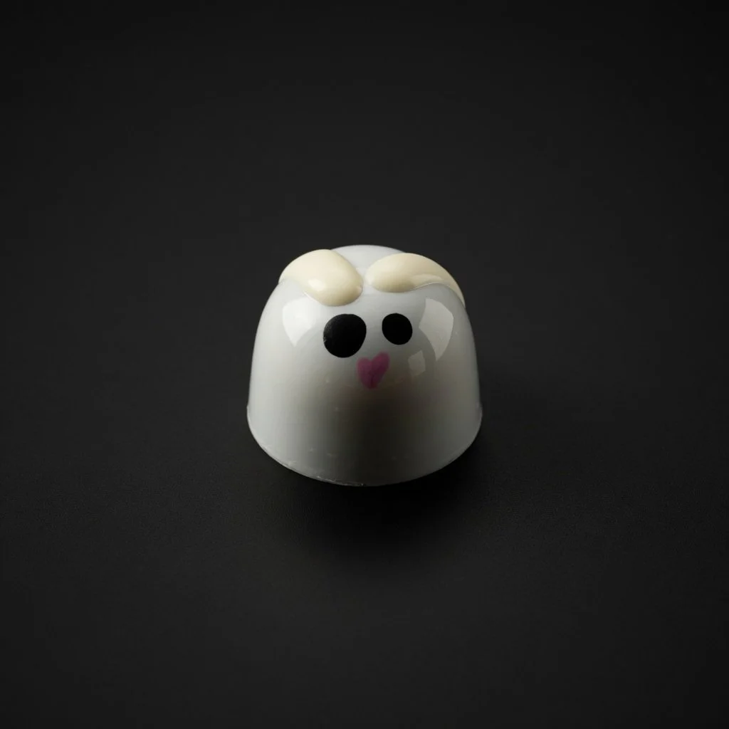 A glossy white chocolate bonbon decorated as a cute bunny face with beige ears, black eyes, and pink heart mouth, sharply in focus and well lit, resting on a solid matte black background with realistic contact shadow.