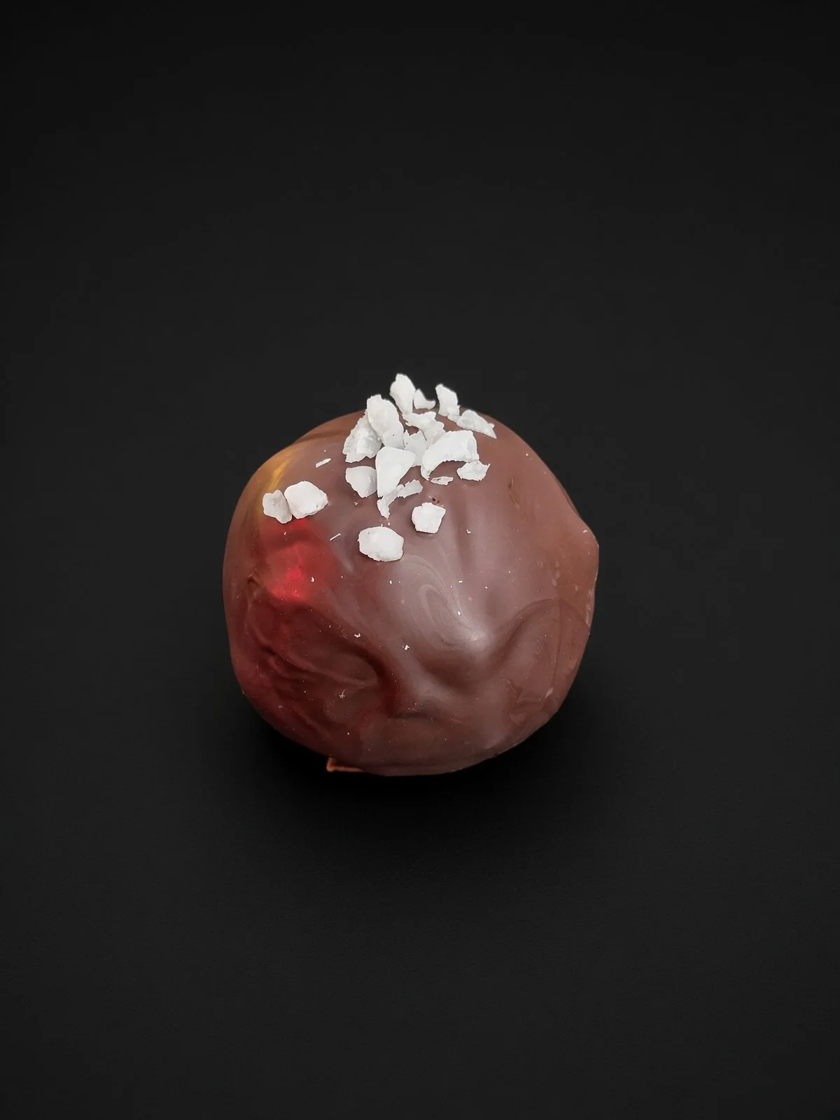 A round chocolate truffle with a blush of red at its base, topped with white coconut flakes, sharply in focus and well lit, resting on a solid matte black background.