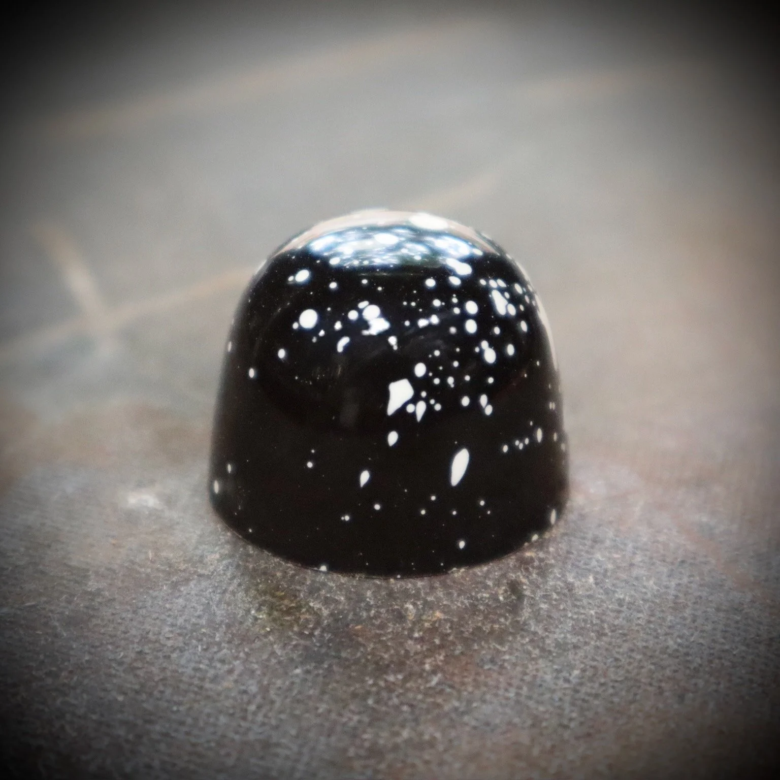 A black, dome-shaped piece of candy with white speckles, placed on a textured surface.