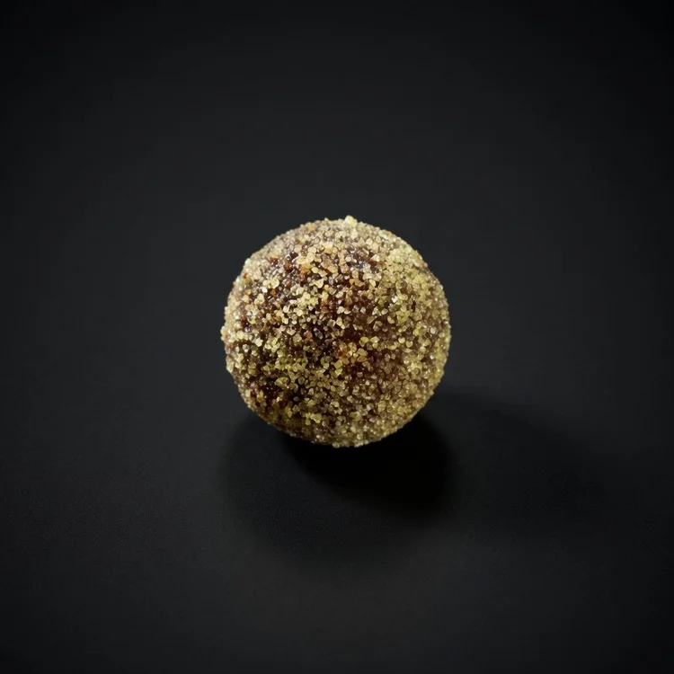 A Passionfruit Explosion Kinetic Bomb: a round chocolate sphere with a crunchy golden-green sugar coating, resting on a solid matte black background.