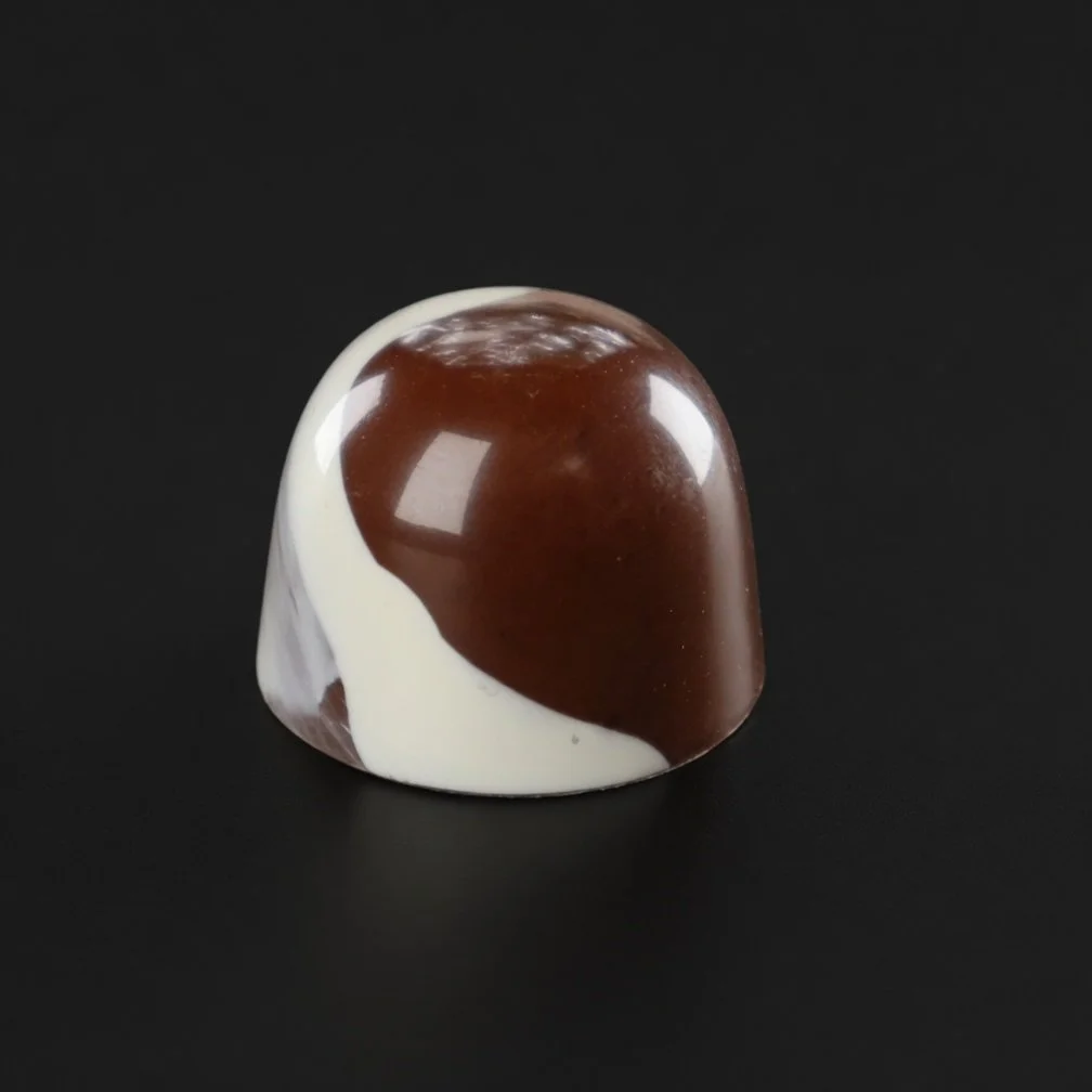 A glossy white-and-brown marbled chocolate bonbon with smooth two-tone swirl, sharply in focus and well lit, resting on a solid matte black background with realistic contact shadow.