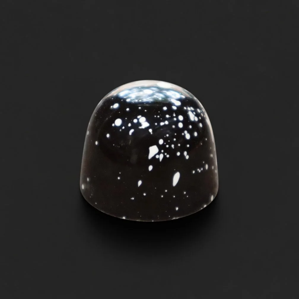 A glossy black chocolate bonbon with white speckles, sharply in focus and well lit, resting on a solid matte black background with realistic contact shadow.