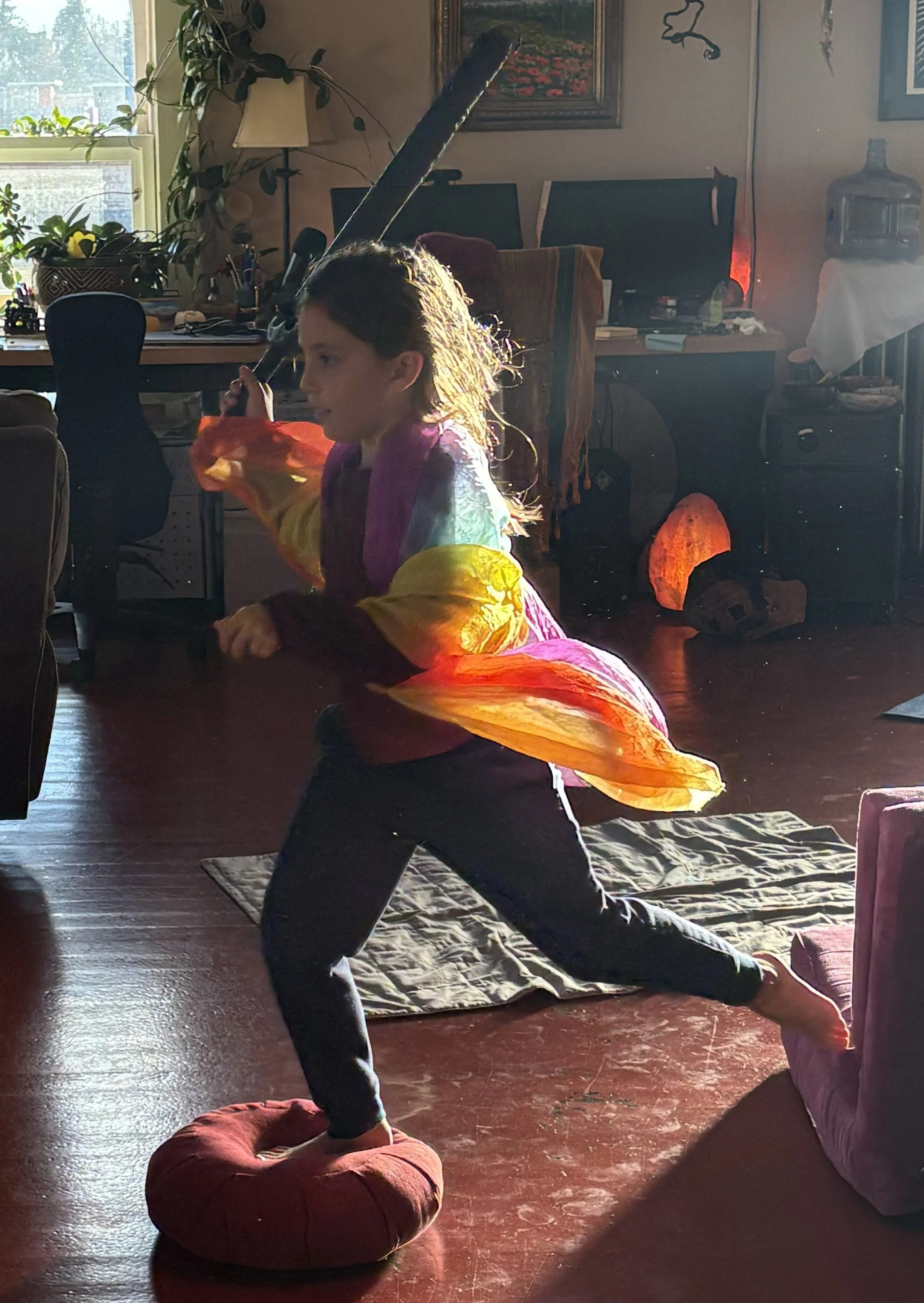 A person in a colorful jacket practicing yoga or stretching indoors on a red cushion, with sunlight streaming through the window illuminating the room decorated with plants, paintings, and furniture.