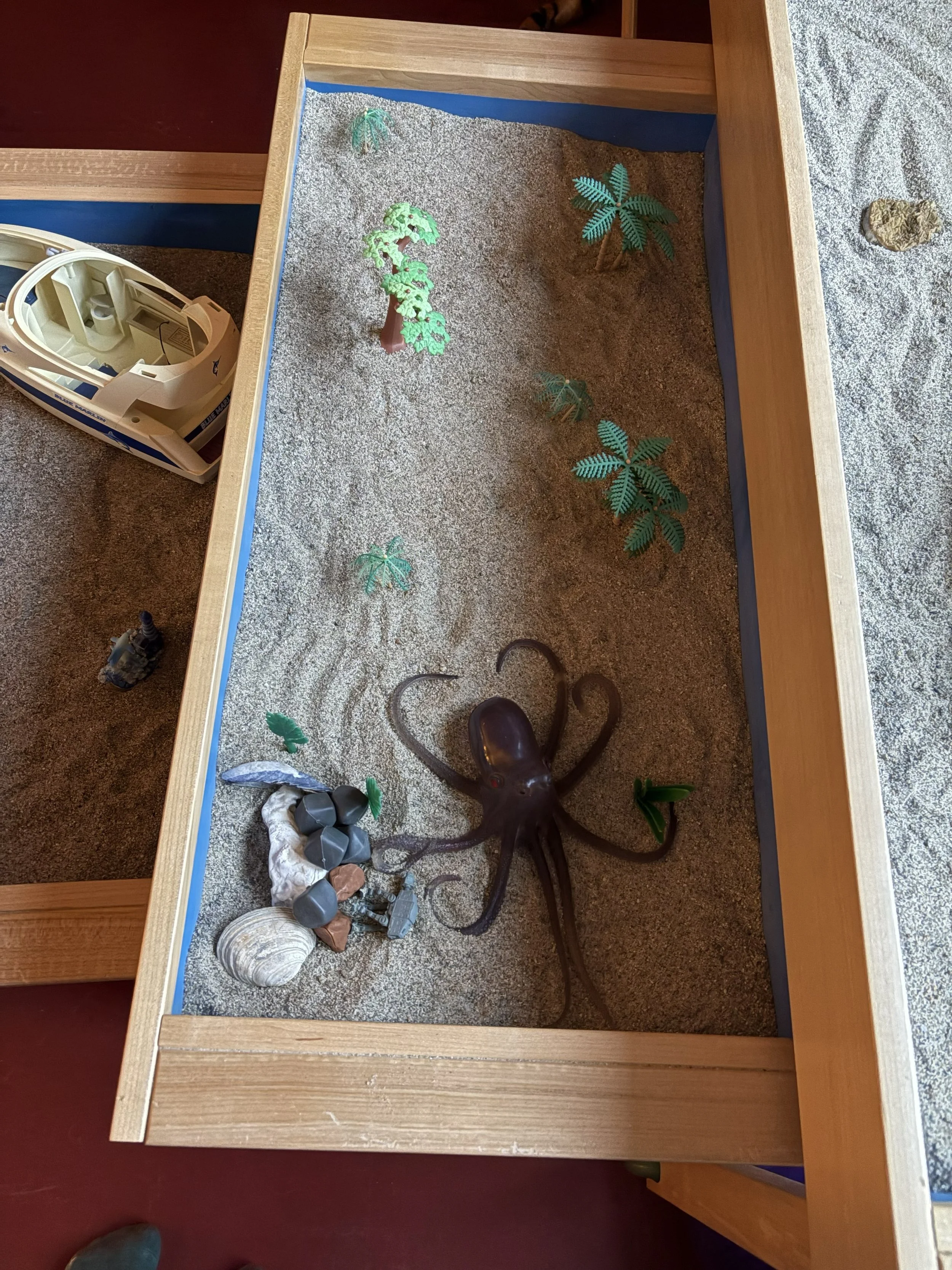 A small indoor desert scene with sand, plastic palm trees, rocks, seashells, and a plastic octopus, inside a wooden frame.