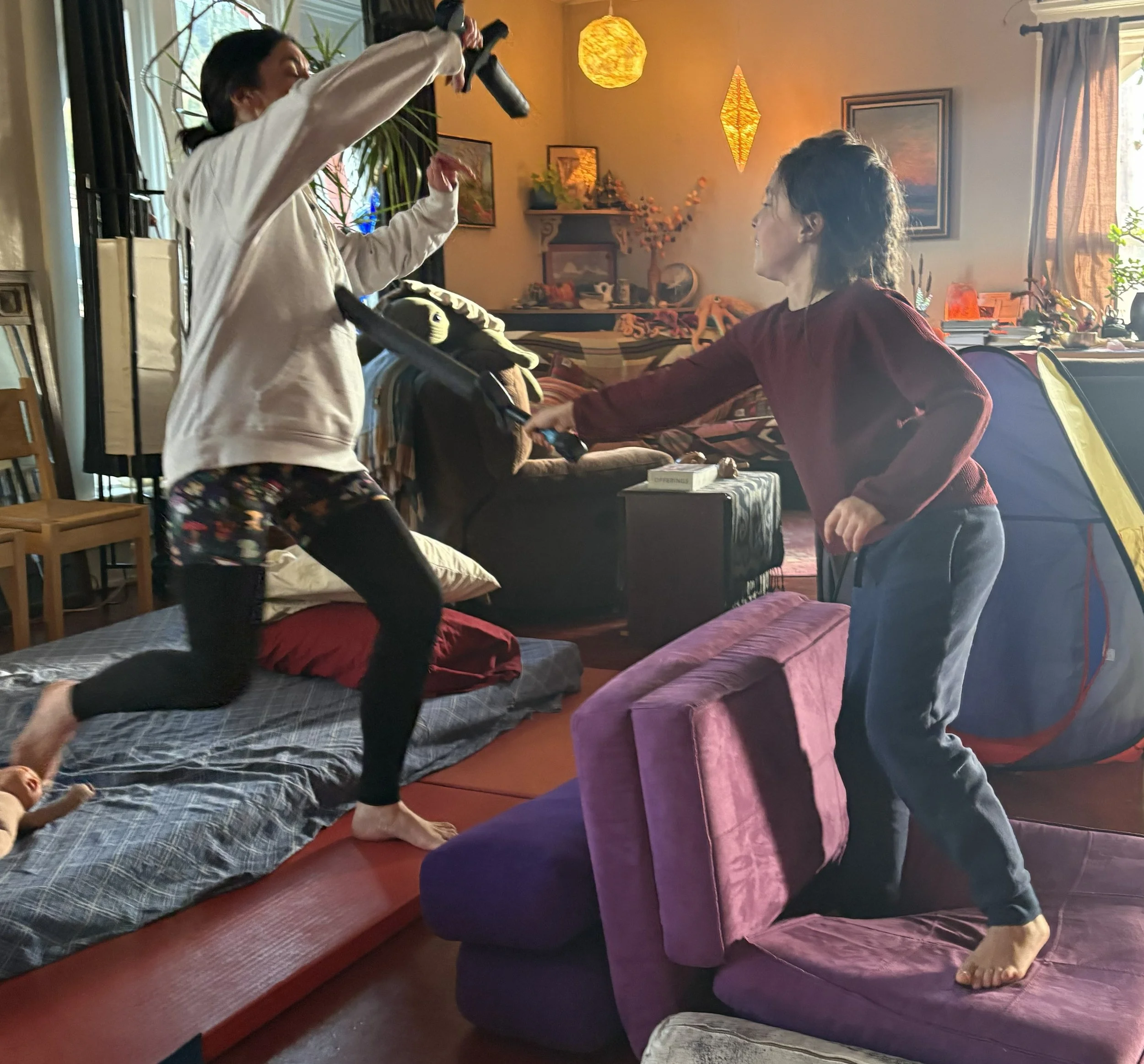 Two women practicing yoga in a cozy living room with warm lighting, decorative lanterns, and plants. One woman is kneeling on a bed with her hands raised, while the other is seated on a chair.