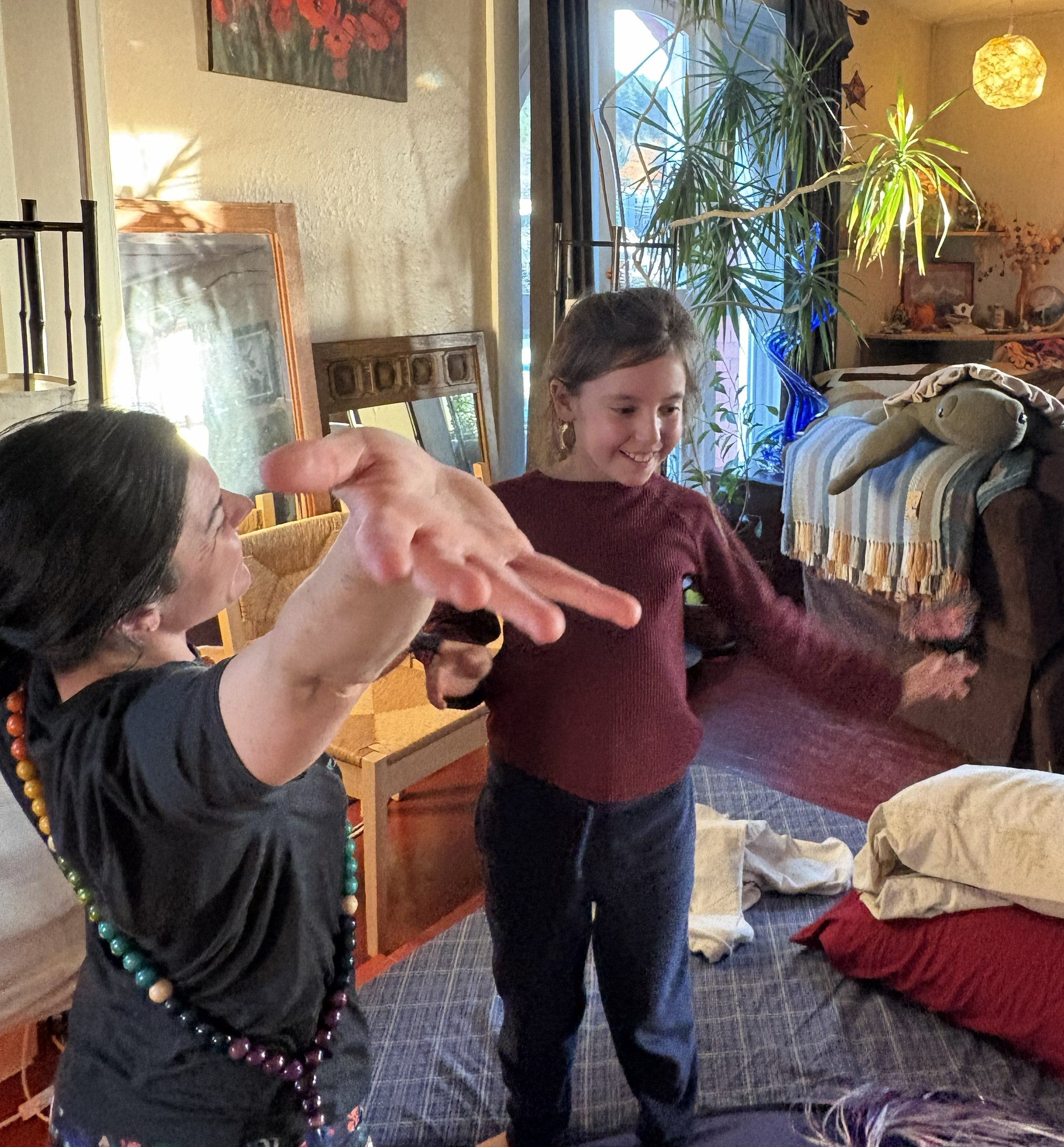 Several people are gathered indoors, engaged in conversation or activities. One woman is wearing a black shirt and colorful beaded necklaces, another person in a maroon sweater is smiling, and another person is wearing a gray shirt and red pants. The room is decorated with houseplants, paintings, and warm lighting.