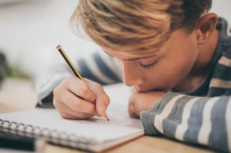 Close-up of a young male student focused on writing in a notebook with a pencil at a school desk, representing campus safety and fire extinguisher inspection education Denver services.