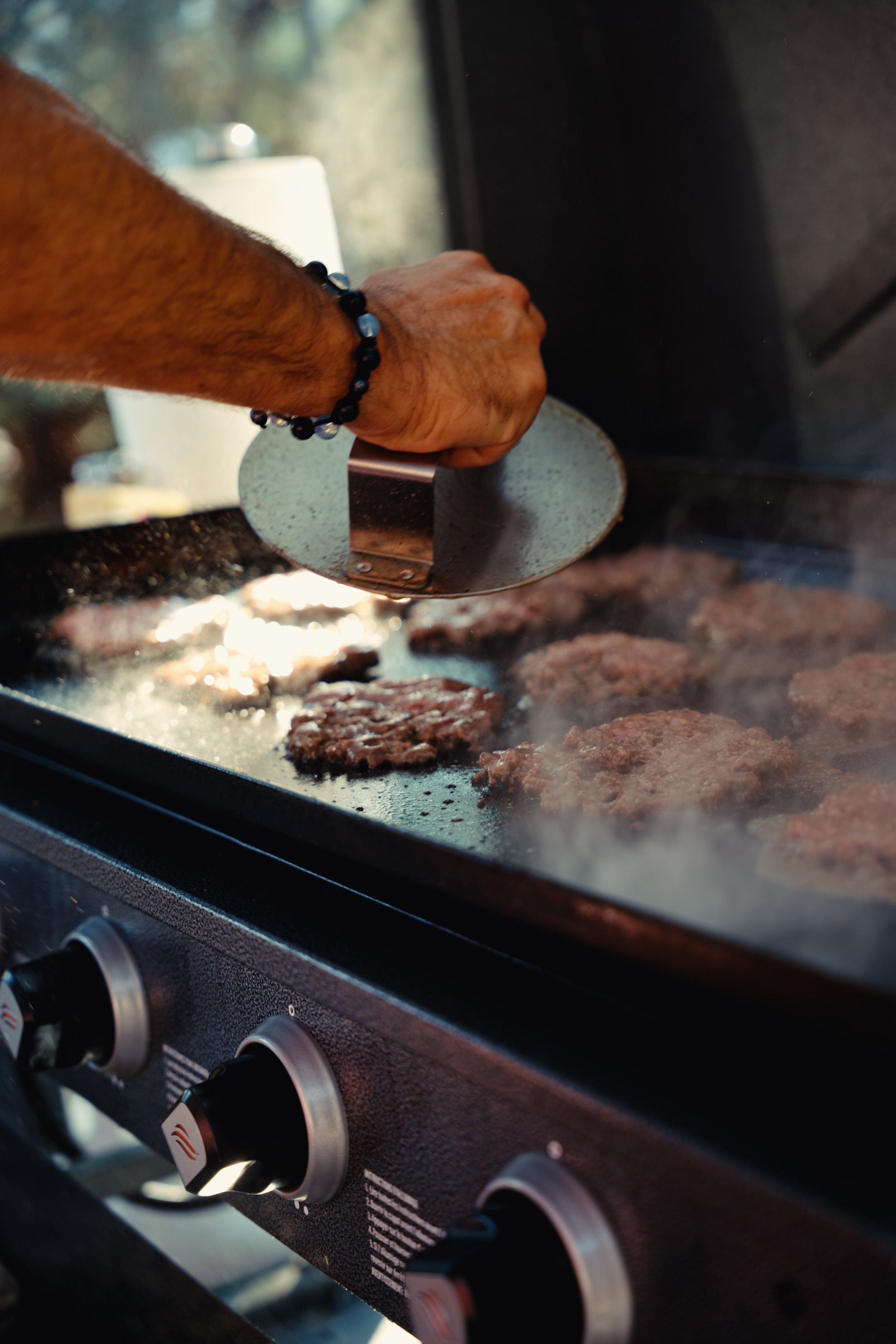 Close-up of a hand holding a spatula, cooking hamburger patties on a stove with a glass door, with visible knobs on the front of the stove.