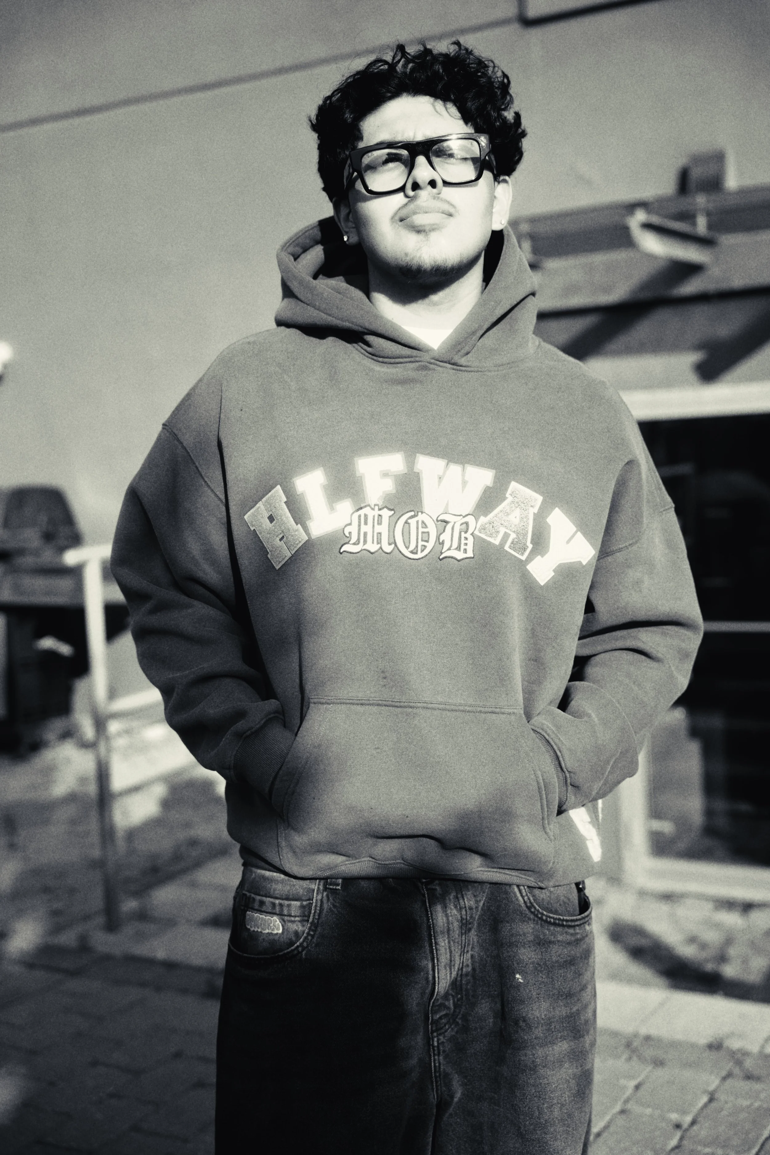 A young man with curly hair, wearing large glasses, a hoodie, and jeans, standing outdoors on a paved area with parked cars in the background.