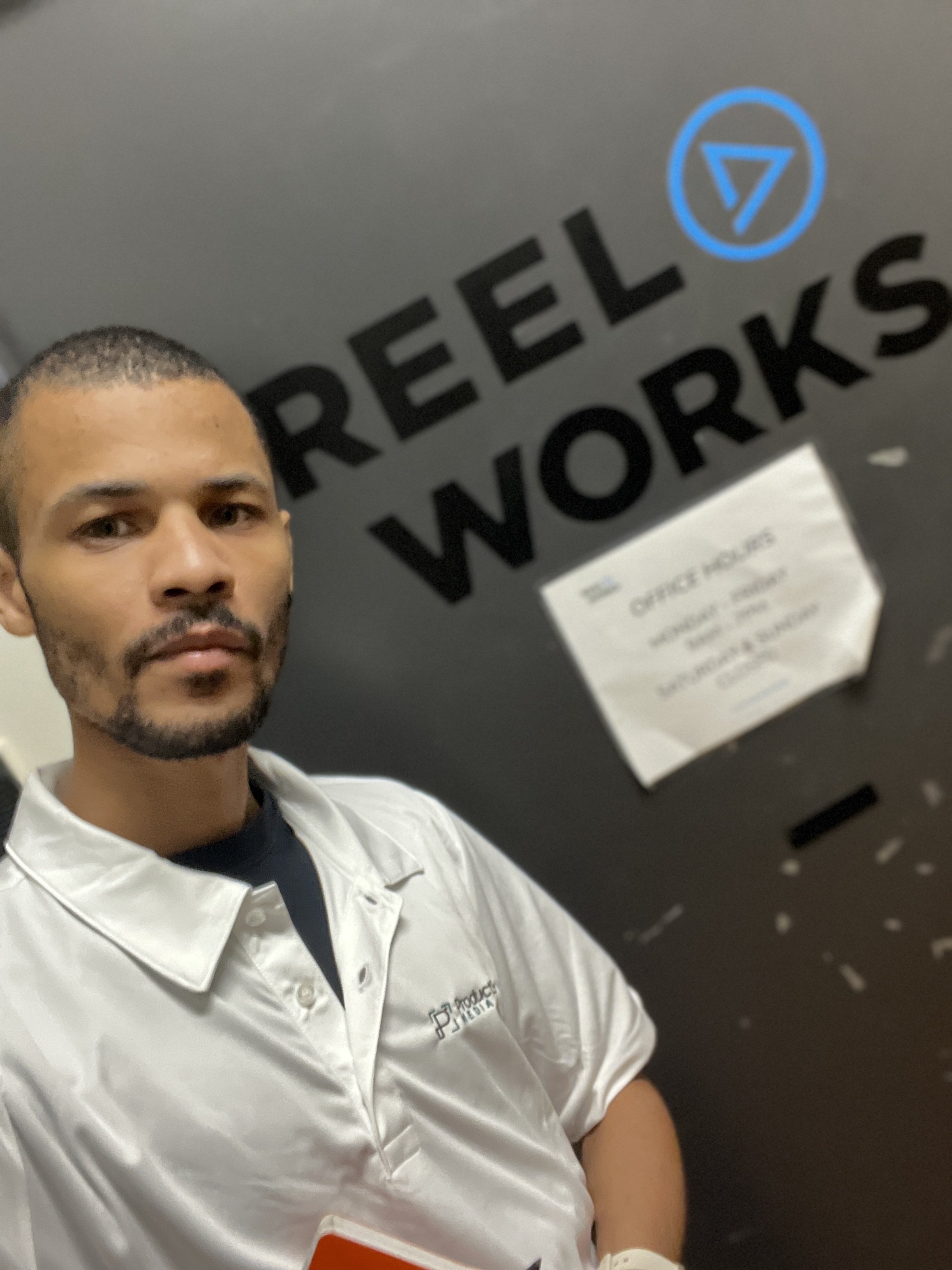 A man taking a selfie in front of a door with the words 'REEL WORKS' and a blue emblem. The man is wearing a white shirt with some text and a black shirt underneath.