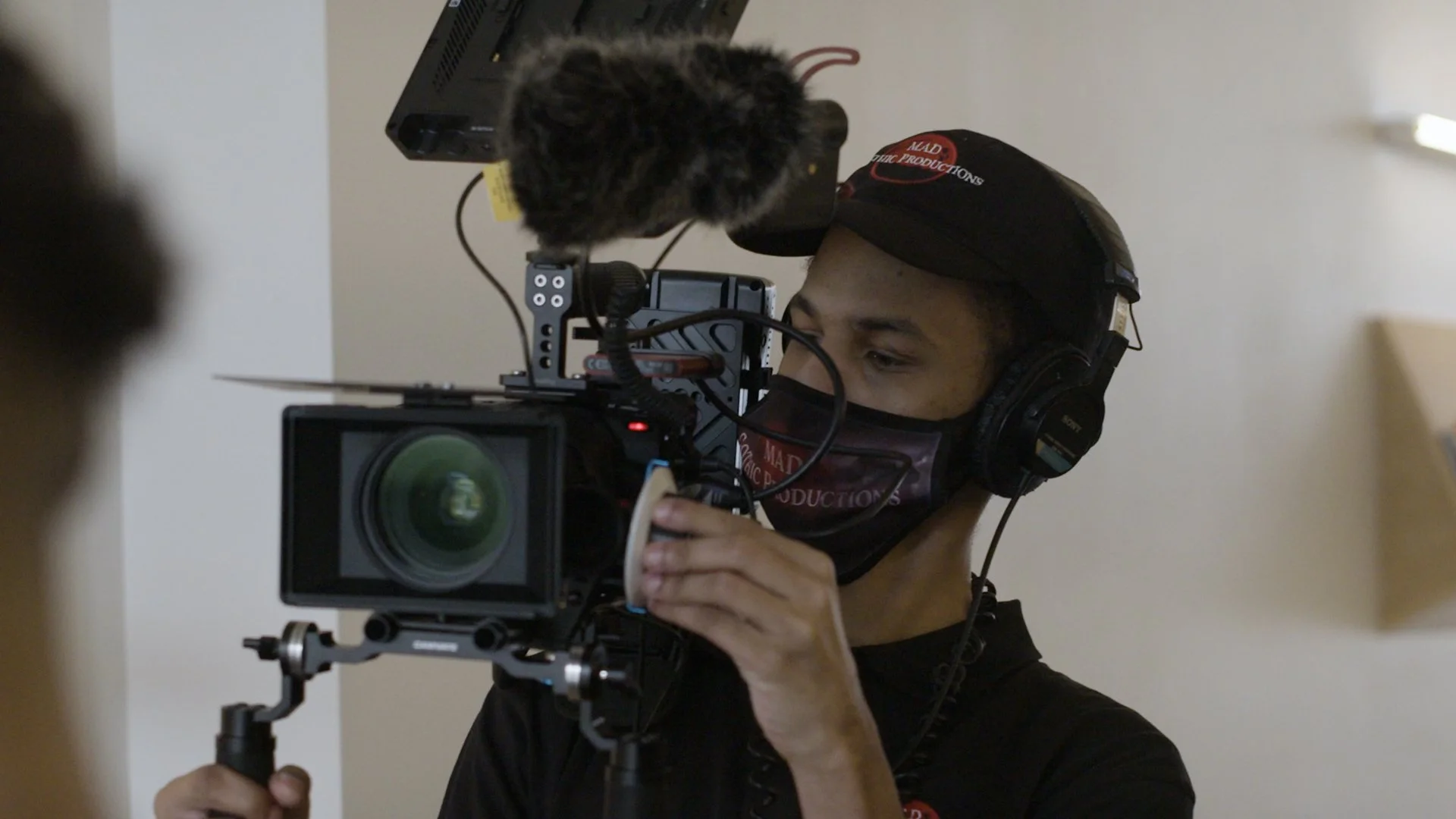 A person wearing a black cap and face mask, operating a professional camera with headphones, in an indoor setting.