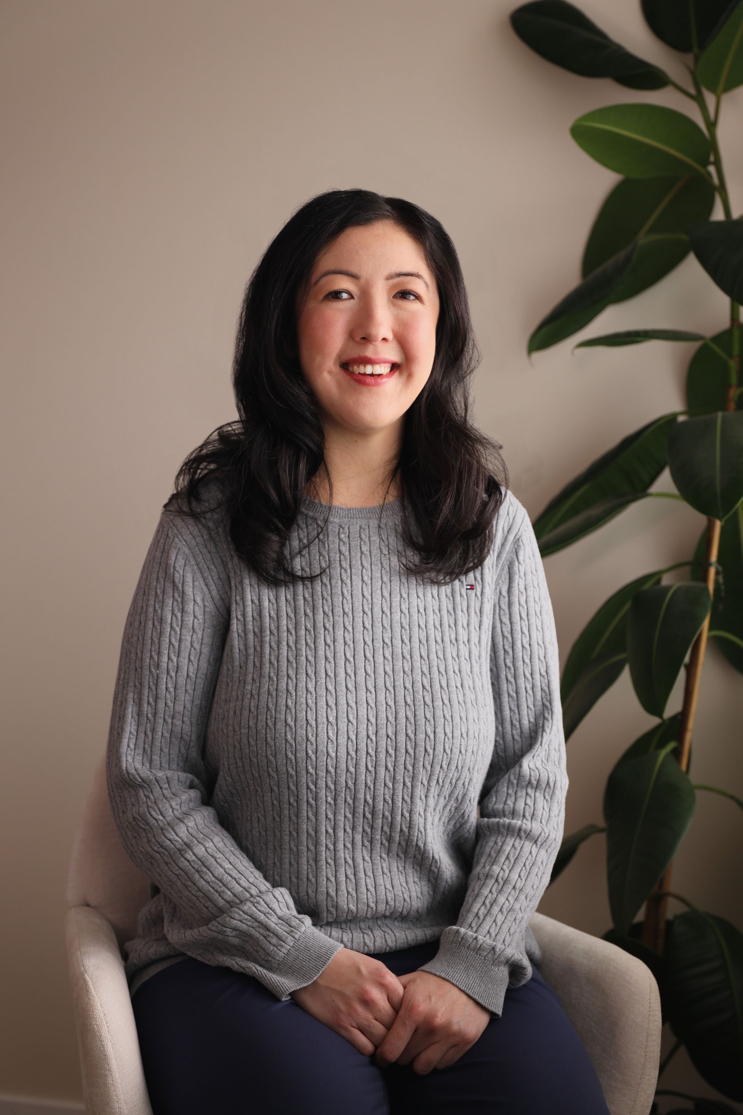 A woman with long black hair smiling while sitting on a beige chair in front of a green plant and a light-colored wall.