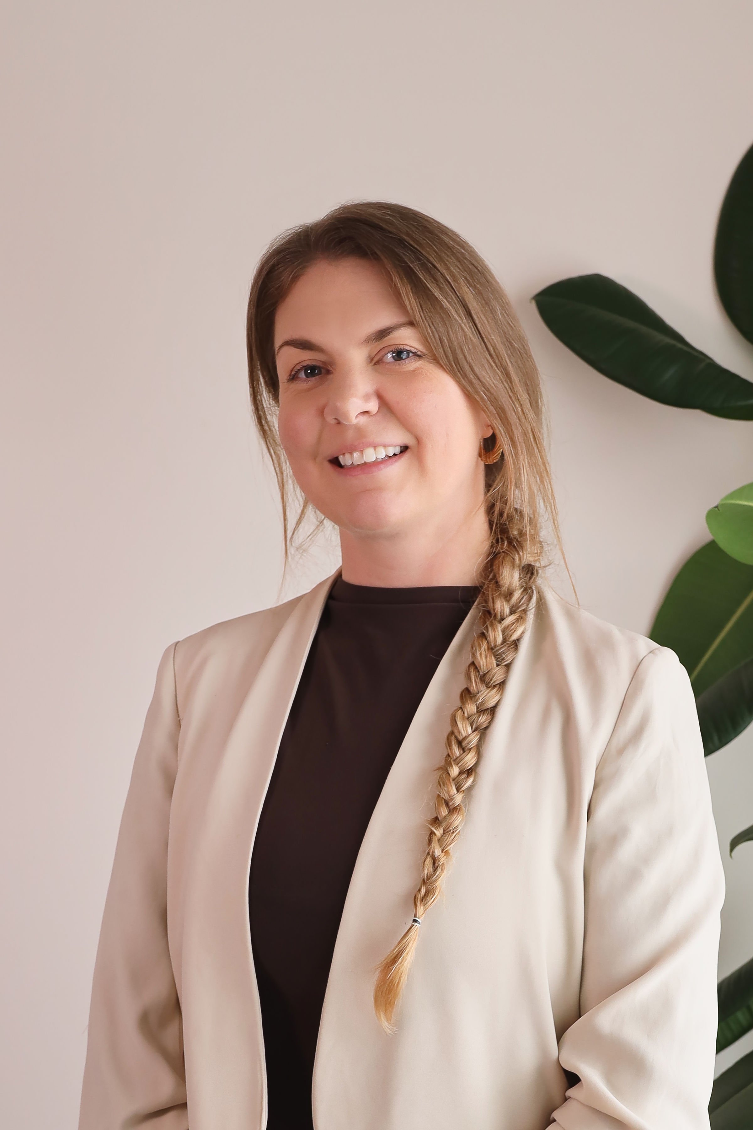 A smiling woman with long, braided hair, wearing a beige blazer over a black top, standing next to a large green plant.