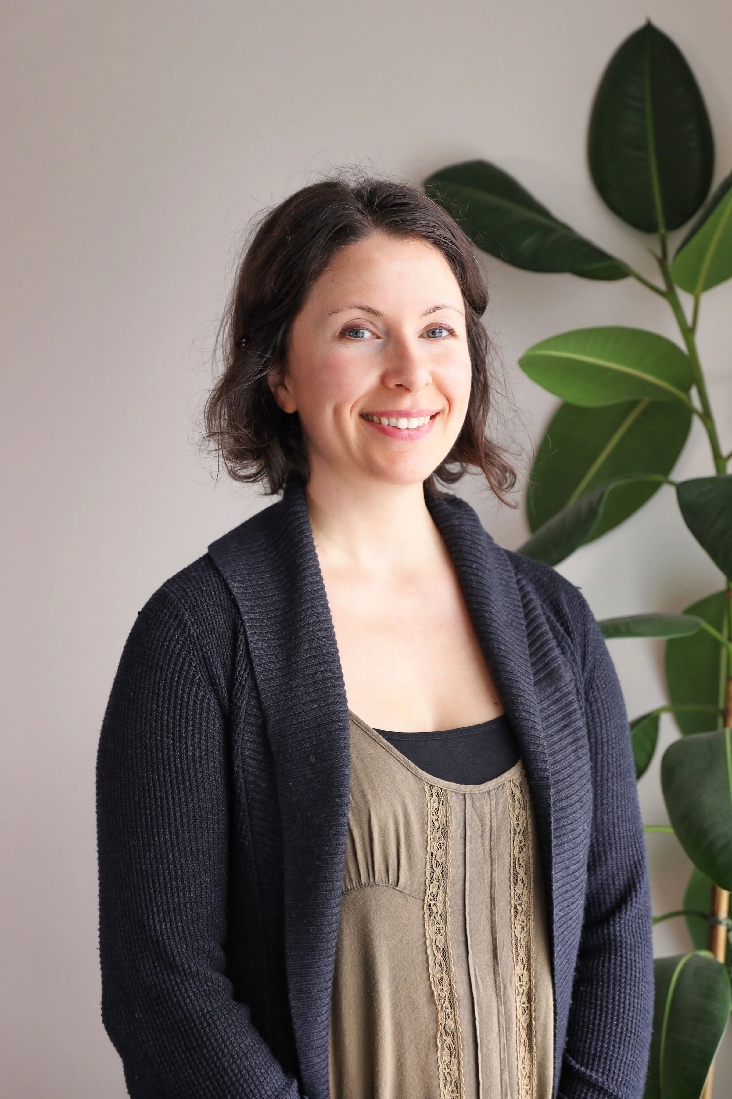 A smiling young woman with dark brown hair in a shoulder-length bob, wearing a black cardigan over a beige top with lace details, standing indoors near a large plant with broad green leaves.