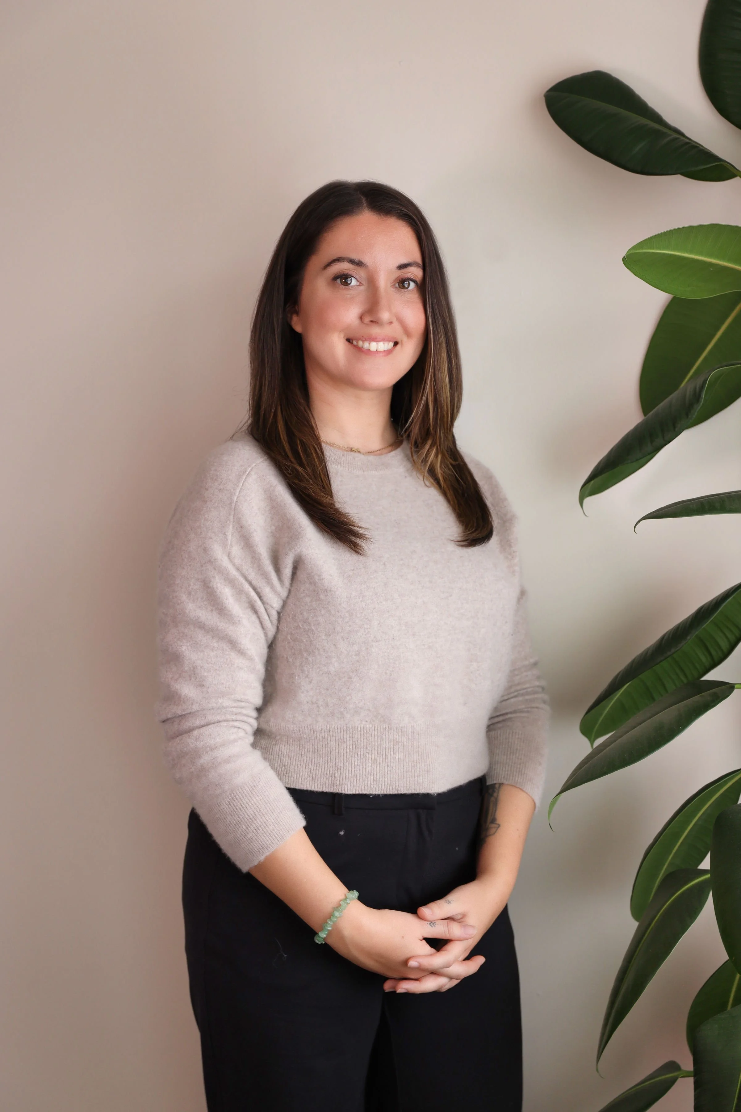 A woman with brown hair and light skin standing indoors against a white wall with a large green plant on her right side. She is smiling, wearing a beige sweater, black pants, and a green beaded bracelet.