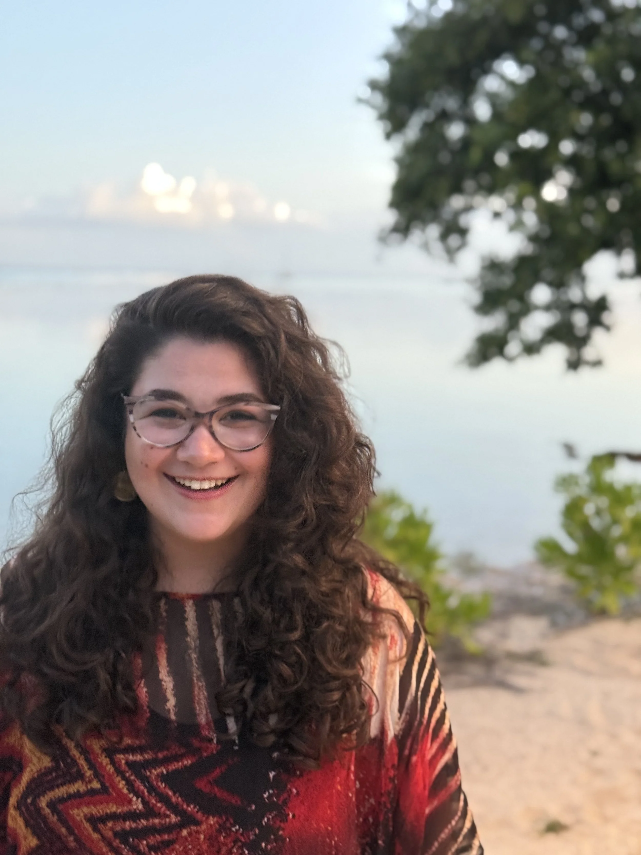 A woman with curly hair and glasses smiling outdoors with trees and a body of water in the background.