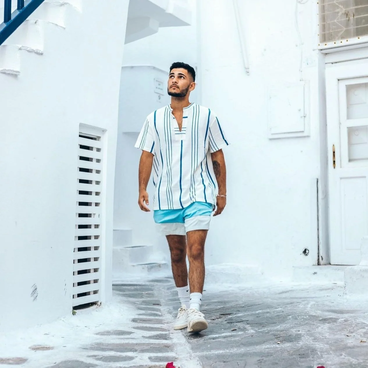 A man walking down a narrow white street, wearing a striped shirt, shorts, and sneakers, with a contemplative expression.