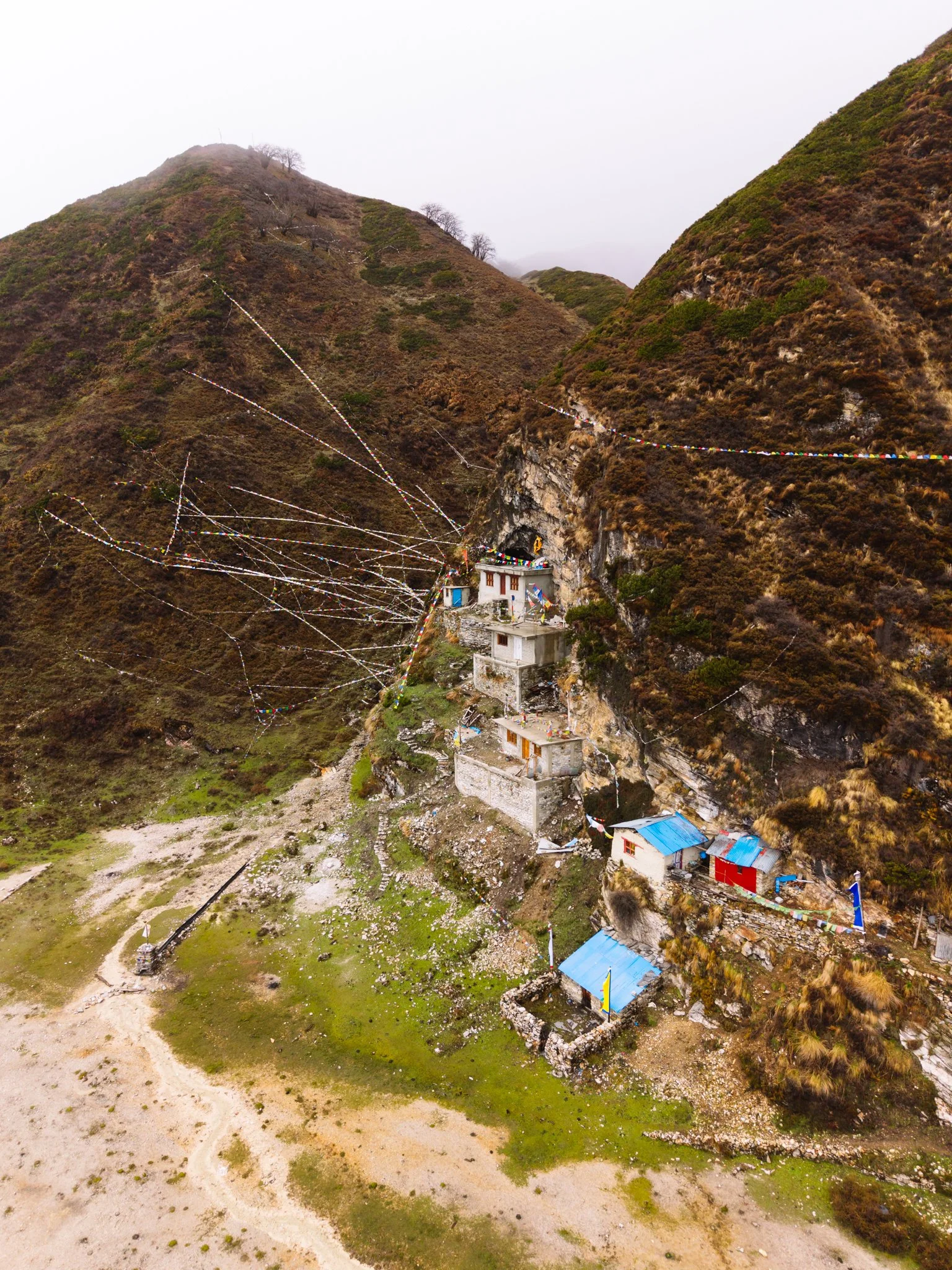 Drone aerial view of Pungyen Gompa monastery built into a cliff with Tibetan prayer flags radiating like a spider web from the surrounding peaks on the Manaslu Circuit Trek