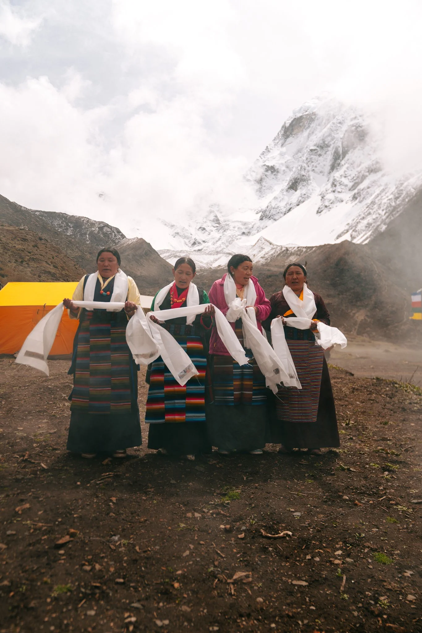Four older Tibetan women in traditional dress performing a khata blessing ceremony with a massive snow-covered Himalayan peak behind on Saga Dawa morning in Samdo village on the Manaslu Circuit Trek