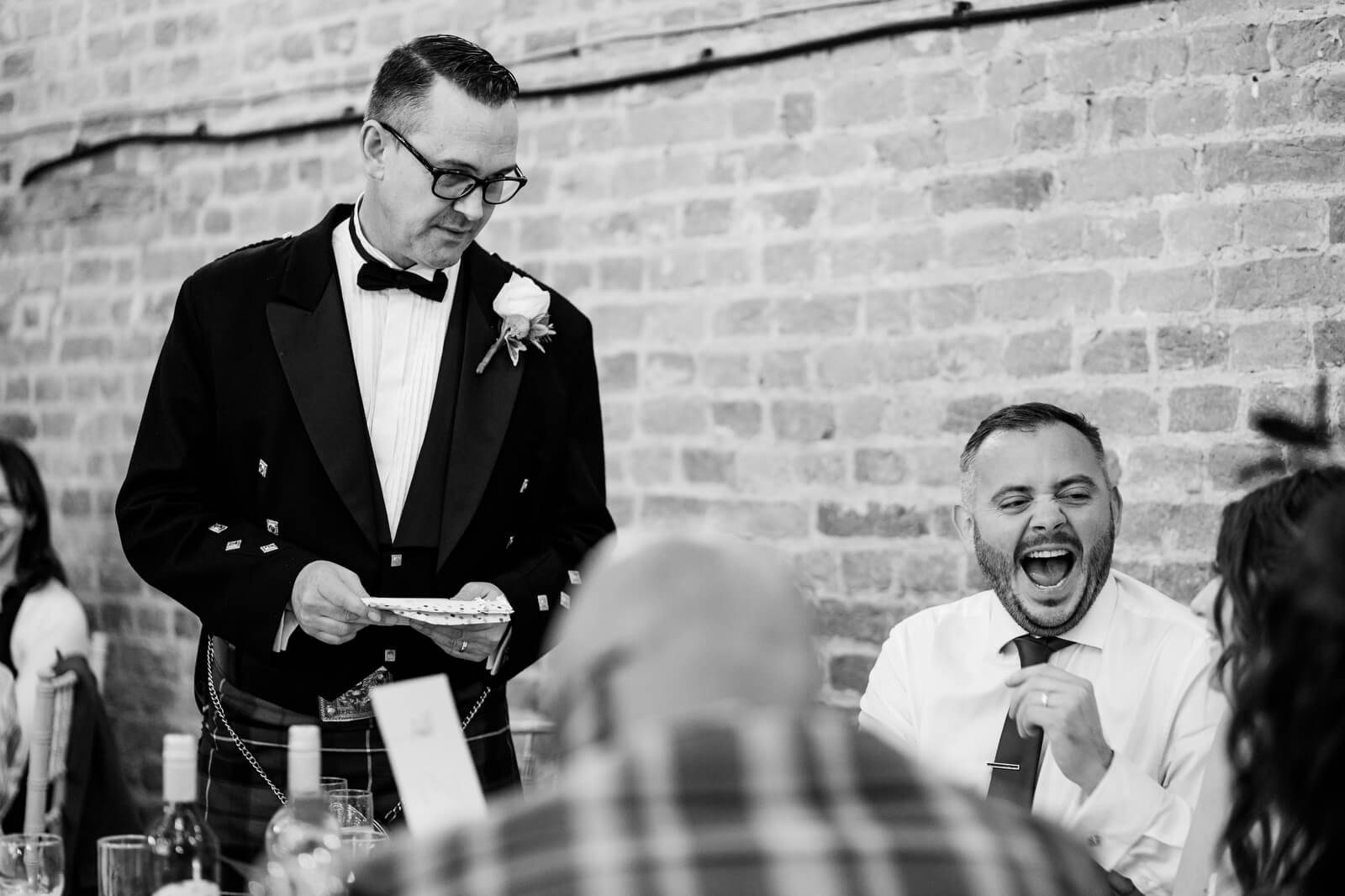 groom laughs during best man's speech at barford park wedding venue in black and white