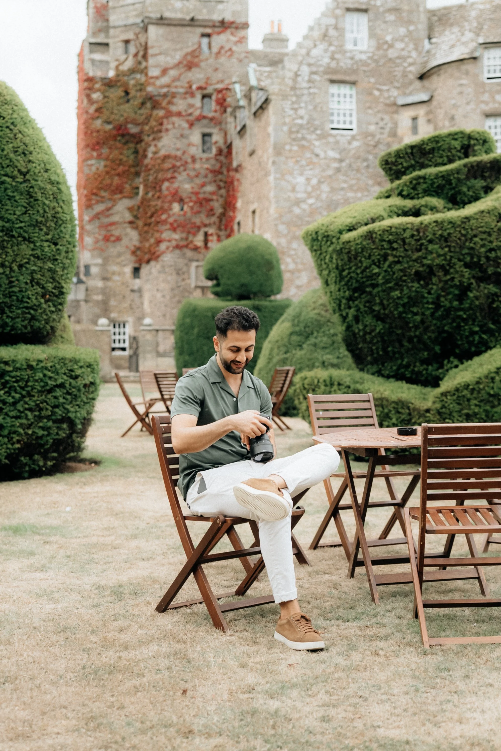 A man sitting on a wooden chair in an outdoor garden area, looking at a camera he is holding, with a castle and well-maintained topiary bushes in the background.