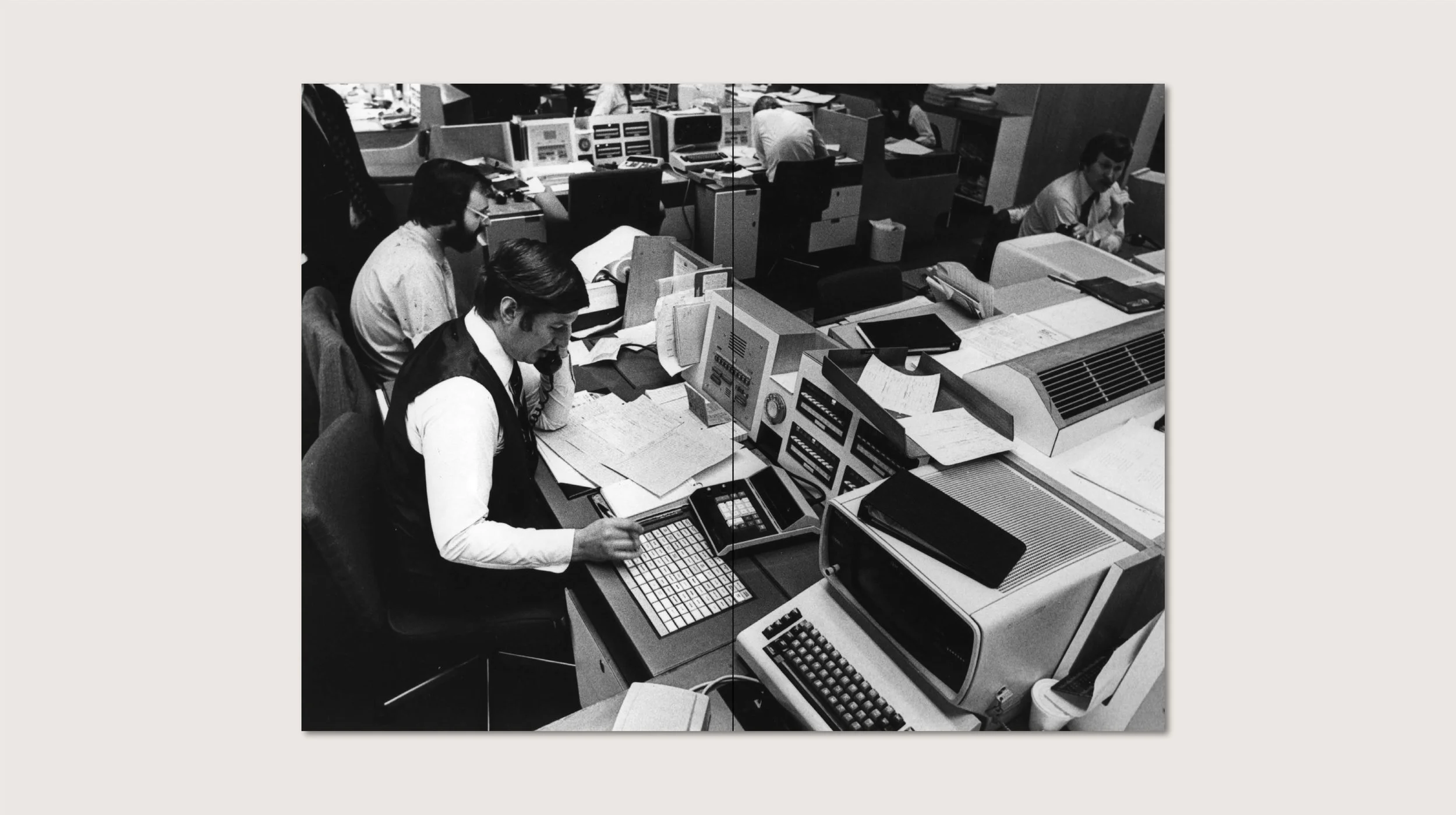 White man sits in an 1970s office surrounded by old technology, writing and on the phone. He is in a small cubicle