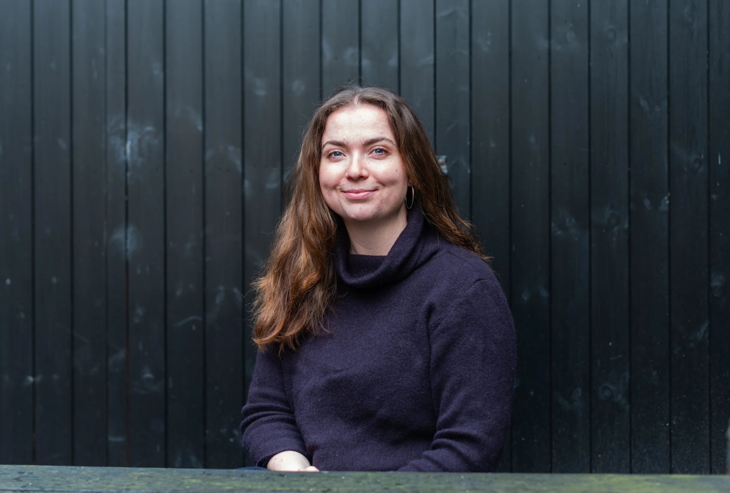 A woman with long brown hair and blue eyes, smiling, wearing a dark sweater, standing against a black wooden fence.