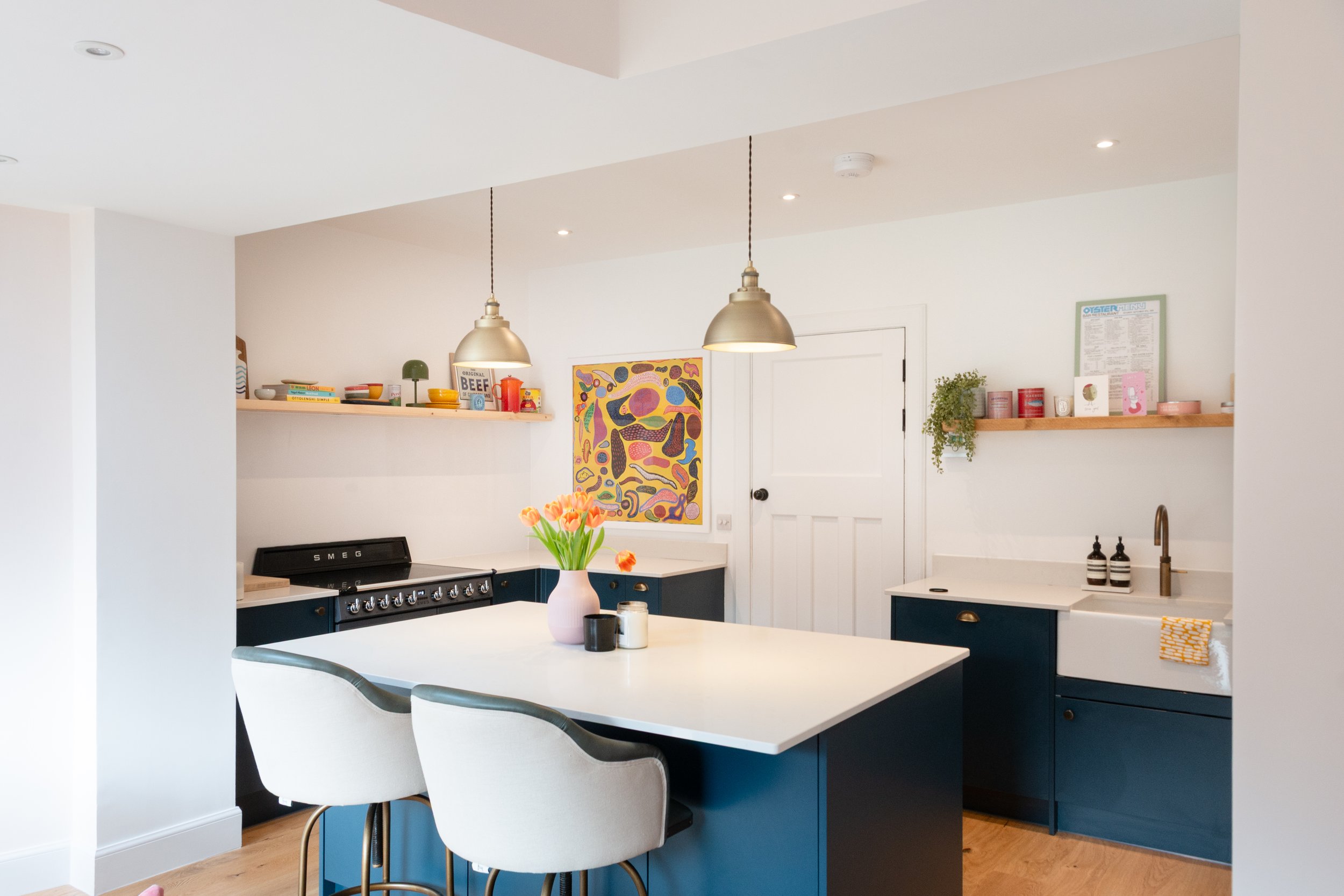 A modern kitchen with white walls and ceiling, recessed lighting, a white kitchen island with a pink vase and flowers, and navy blue cabinets. Open shelving displays colorful dishes, books, and decor. There are pendant lights hanging above the island, a colorful abstract painting on the wall, and a white door.