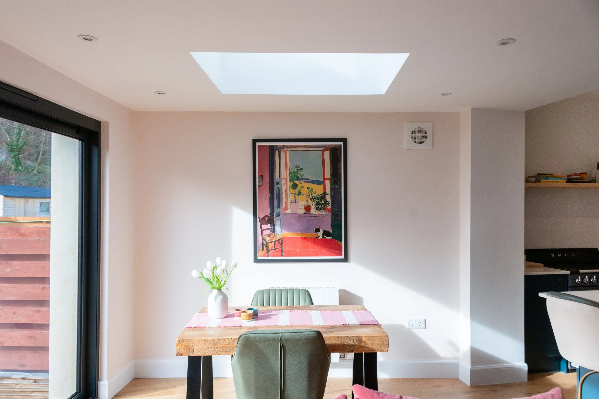 A dining area with a wooden table, green upholstered chair, and a vase with white flowers. A colorful painting hangs on the wall, showing an open window with a view of green plants and a field. There is a large skylight in the ceiling providing natur