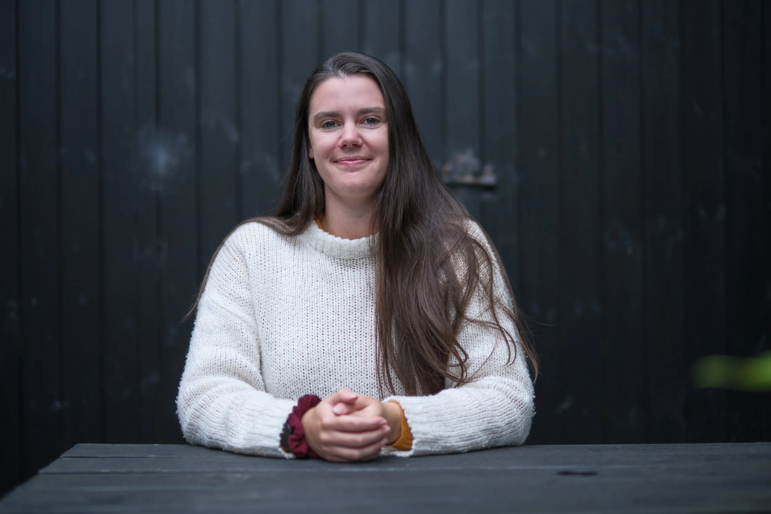 A woman with long brown hair wearing a cream knit sweater sitting at a dark table outdoors, smiling, with a dark wooden fence in the background.