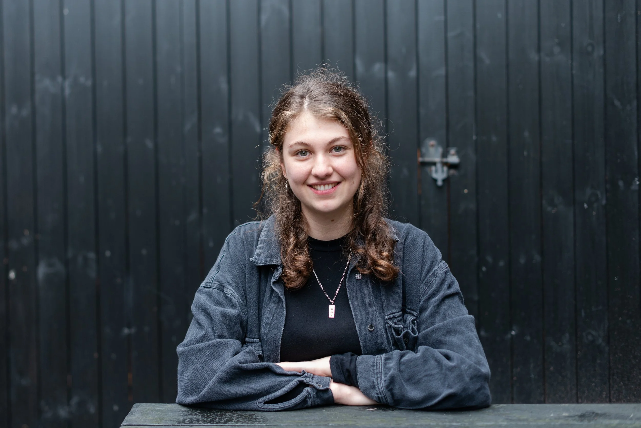 A young woman with long, curly hair wearing a black shirt, a black jacket, and a silver necklace, sitting at a dark wooden table with her arms crossed in front of her, smiling, in front of a black wooden background.