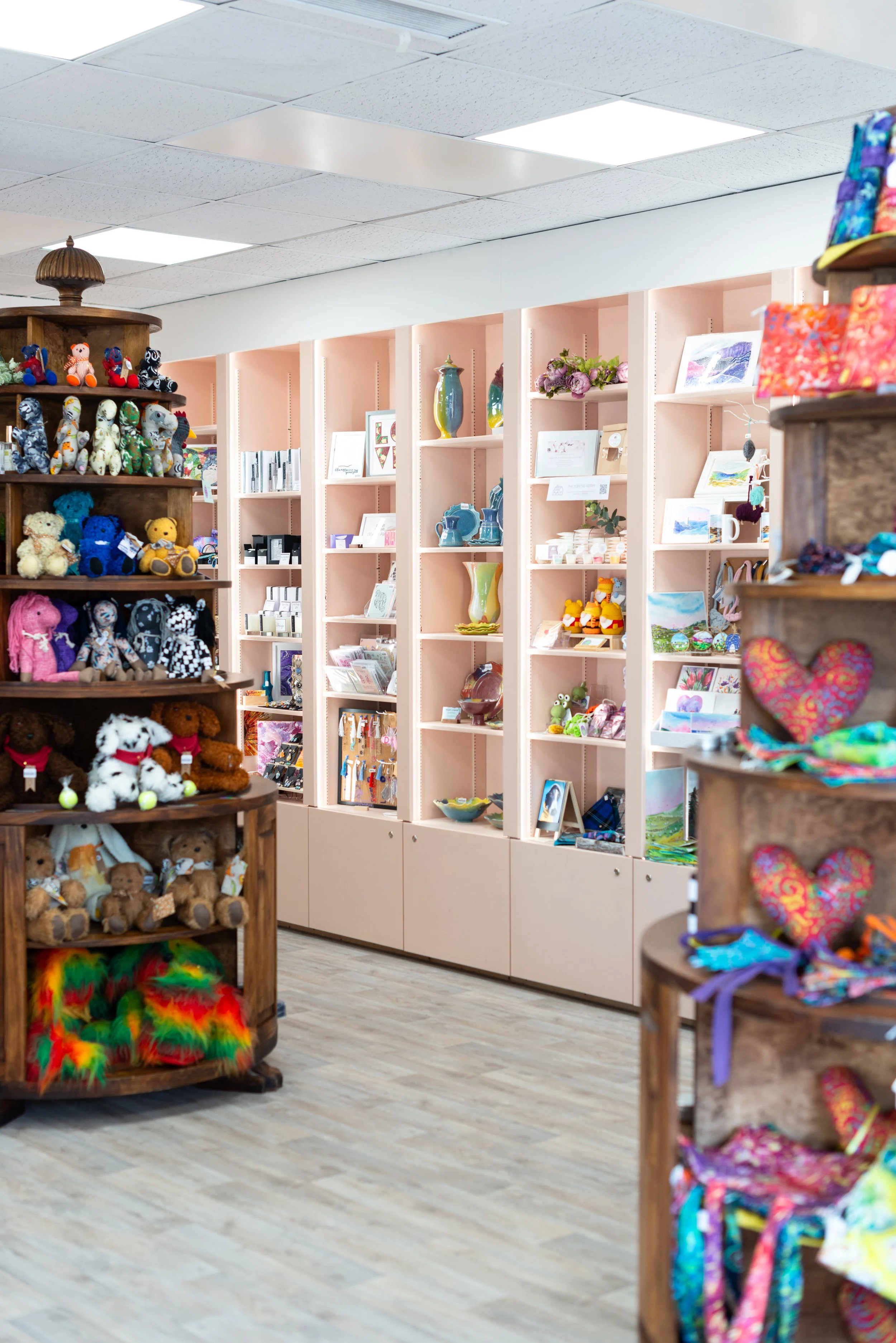 Interior of a gift shop with shelves displaying colorful plush toys, ceramics, framed artwork, and handmade crafts, with a light wood floor and white ceiling tiles.