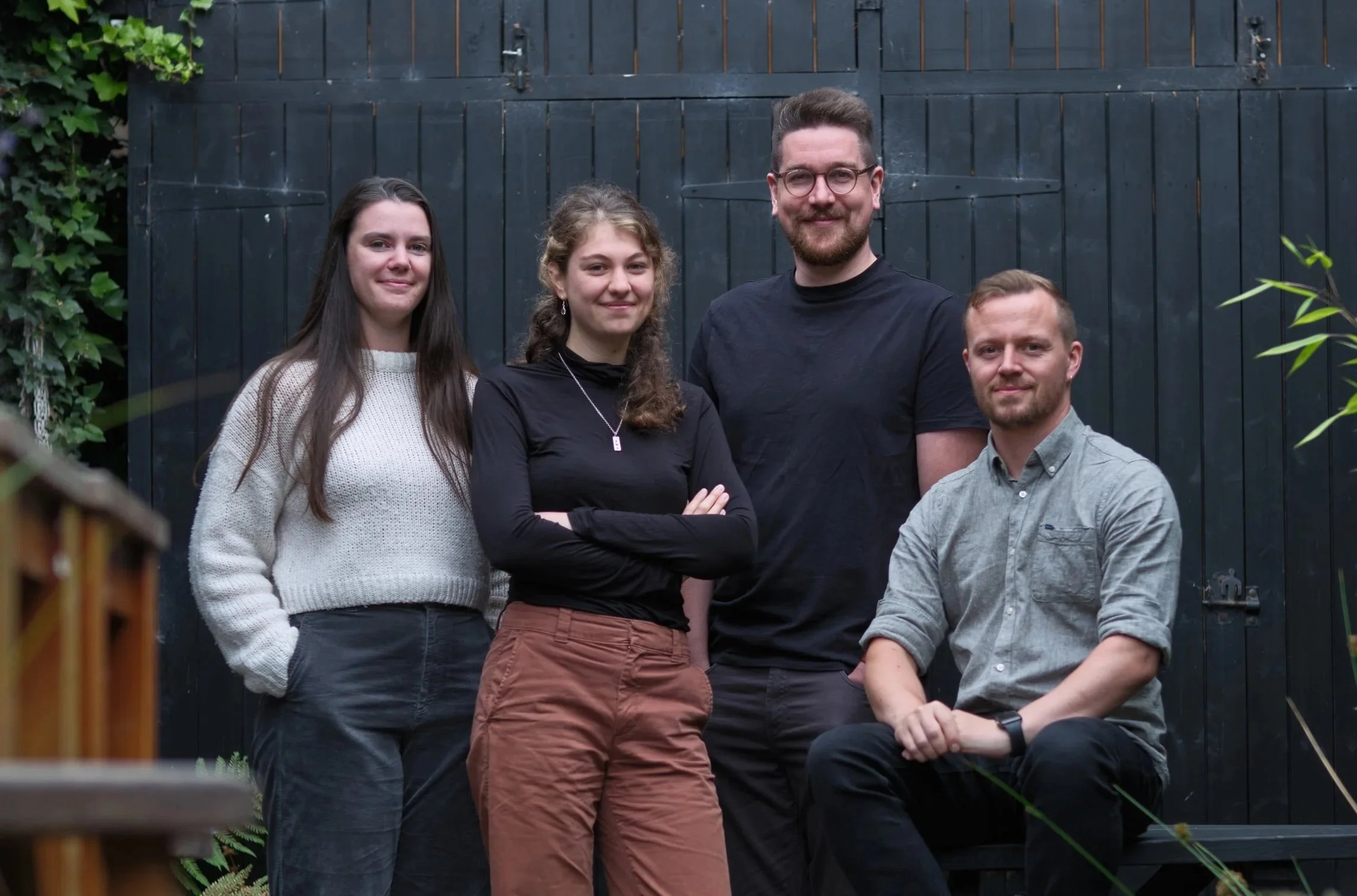 A group of five diverse young adults, three women and two men, standing and sitting outdoors in front of a black wooden fence, smiling and posing for a photo.