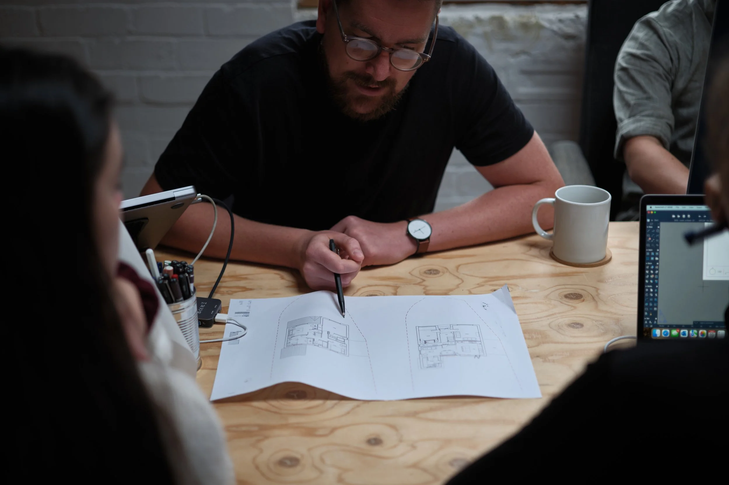 A man with glasses and a beard sketching floor plans on paper with architectural drawings on a wooden table, surrounded by colleagues in an office setting, with a coffee mug and laptop visible.