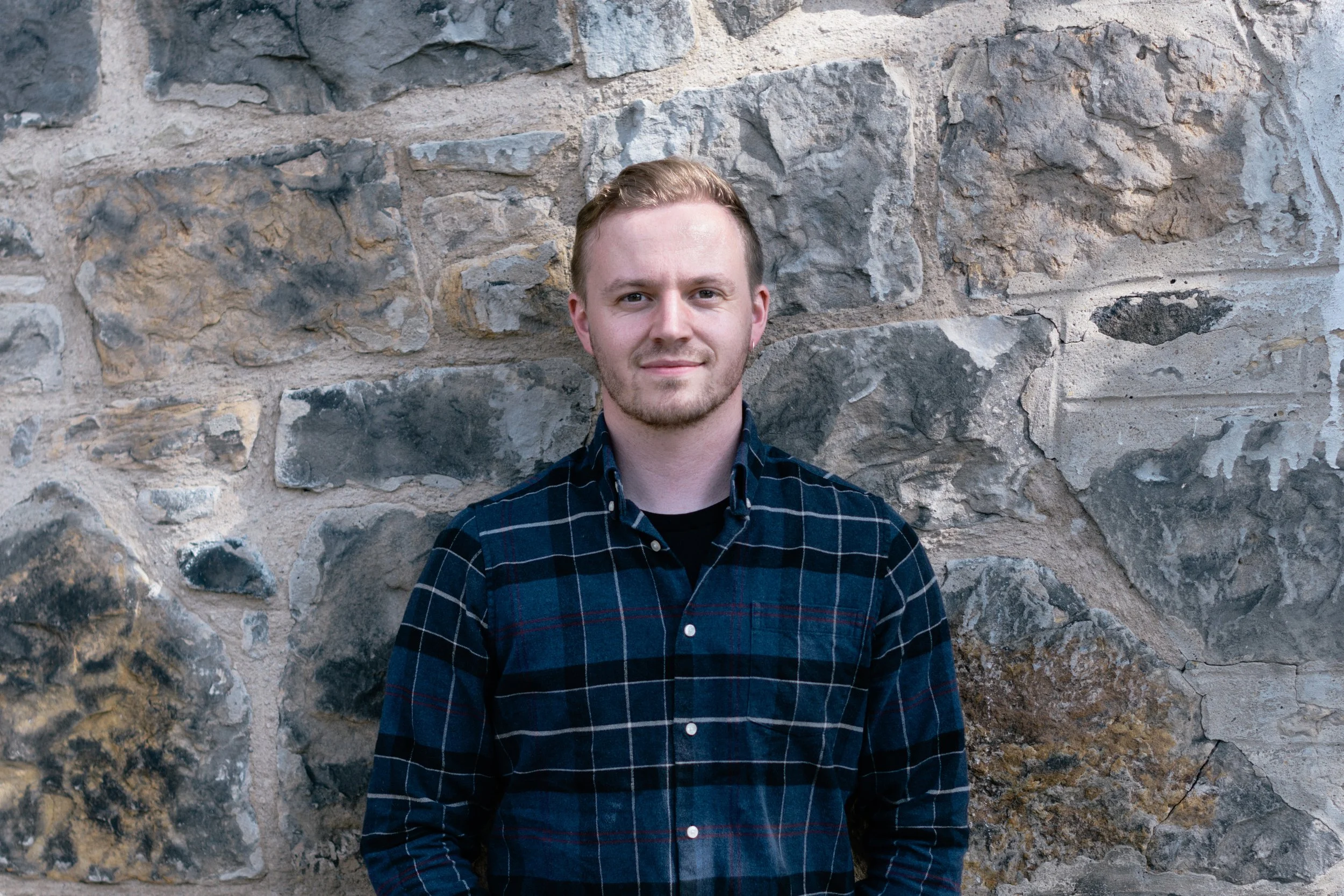 A young man with short hair and light facial hair, wearing a dark plaid shirt, standing in front of a stone wall.